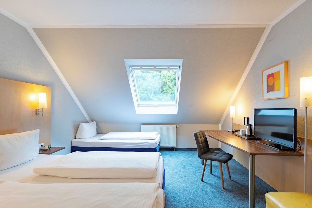 Una camera d'hotel con un letto, una scrivania con un computer, una sedia, una finestra e un quadro appeso al muro.