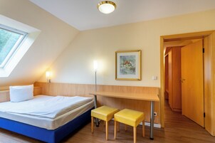 Un dormitorio con una cama, un escritorio, dos taburetes amarillos, un cuadro enmarcado en la pared y una puerta que da a otra habitación.