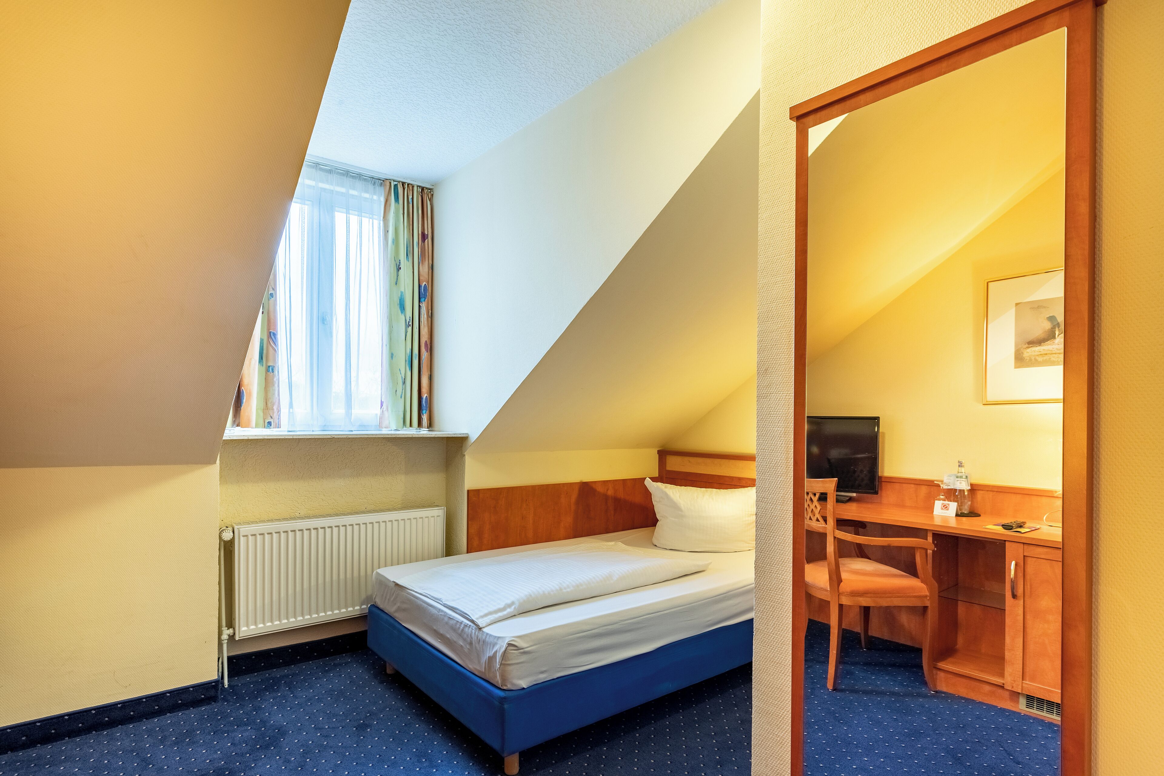 A bedroom with a bed, a desk, a chair, a radiator, and a window with curtains.
