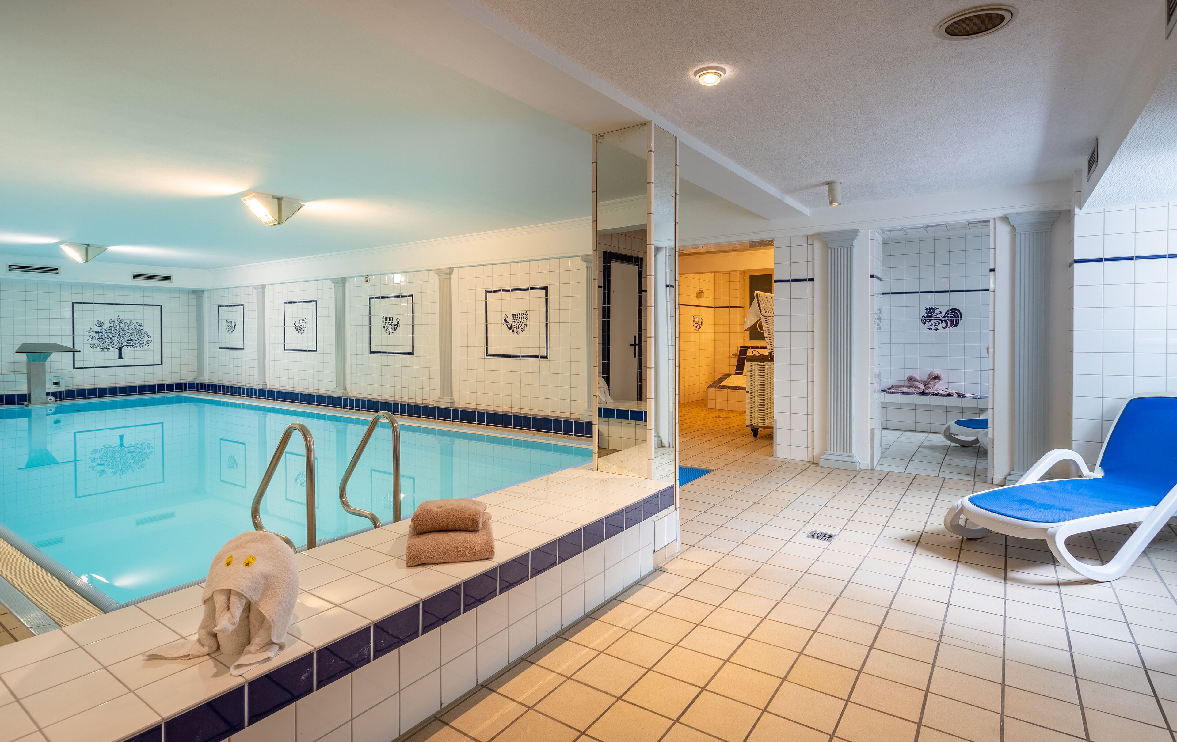 Ein Schwimmbecken mit gefliesten Wänden und blauer Umrandung, ein Handtuchhalter mit Handtüchern sowie ein blauer Liegestuhl.