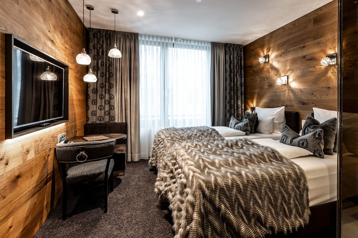 A modern hotel room with a wooden wall, a large bed with patterned bedding, a desk with a chair, and a large window with curtains.
