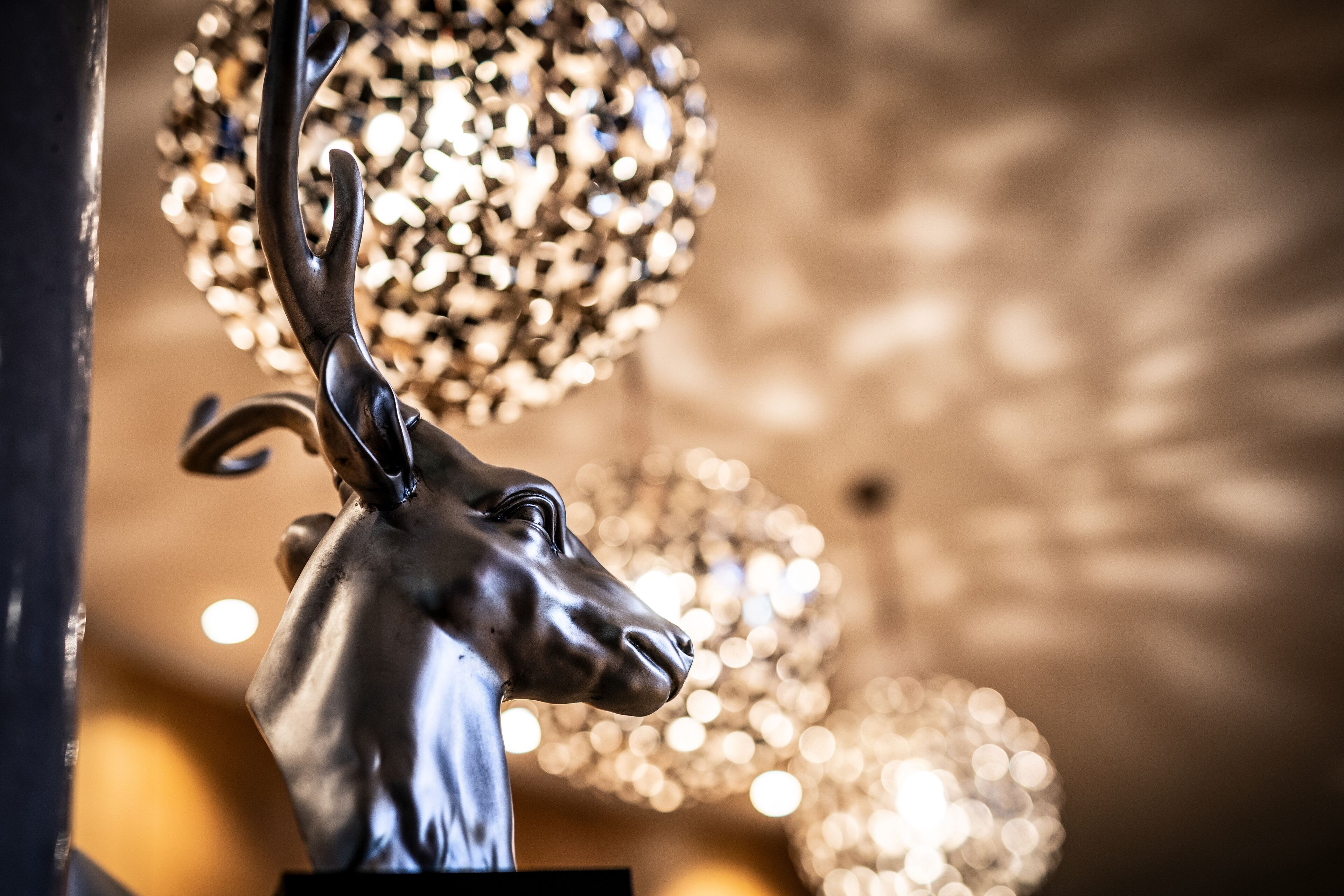 Een close-up van een metalen hertenhoofdbeeld met een wazige achtergrond, waarin een decoratieve lamp te zien is.