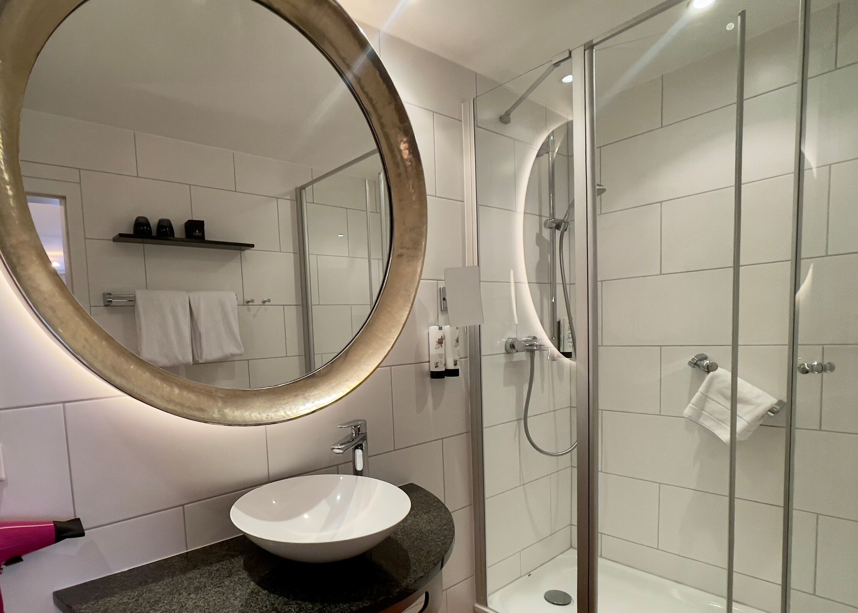 A modern bathroom with a round mirror, a white sink, a glass-enclosed shower, and white tiled walls.