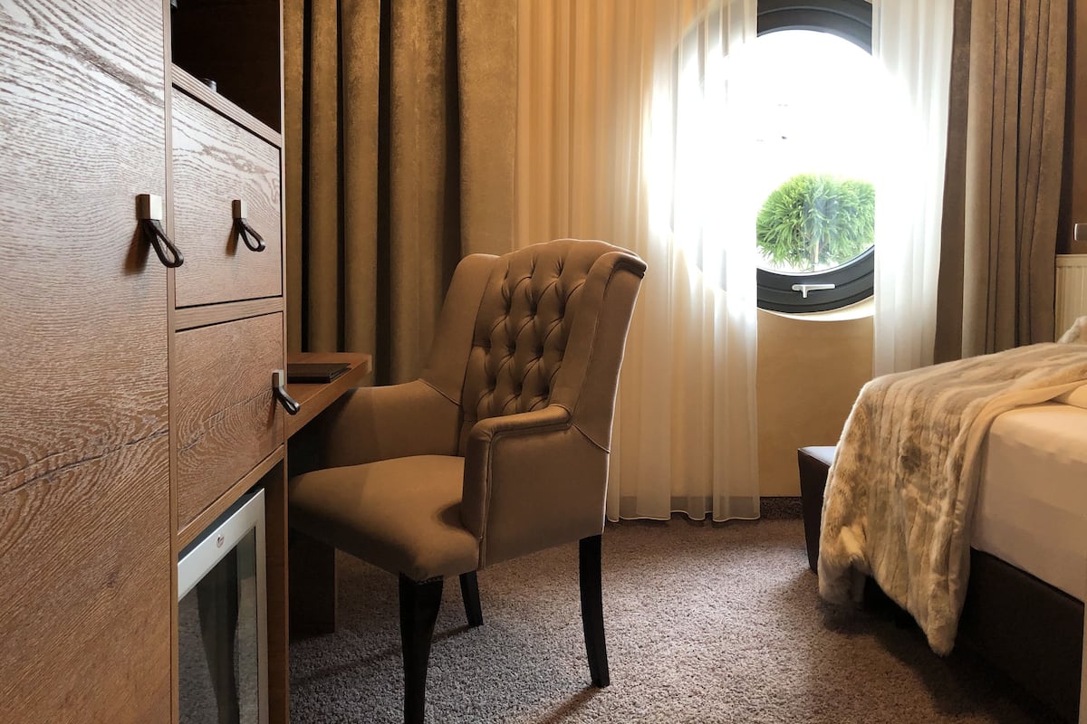 A hotel room with a tufted armchair, a bed with a white blanket, a wooden dresser, and a window with a view of greenery.