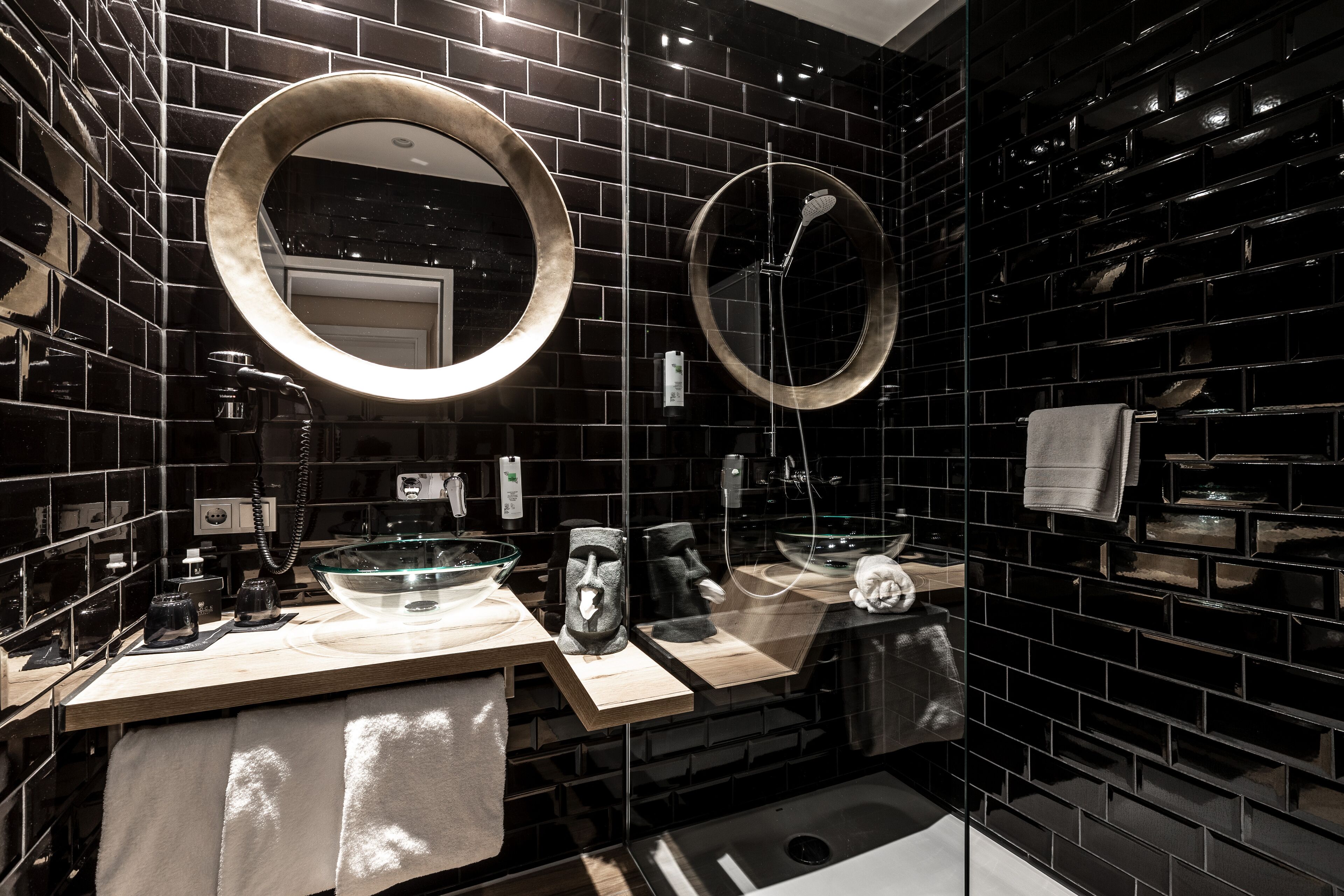 A modern bathroom with black tiled walls, two round mirrors, and a glass bowl sink.