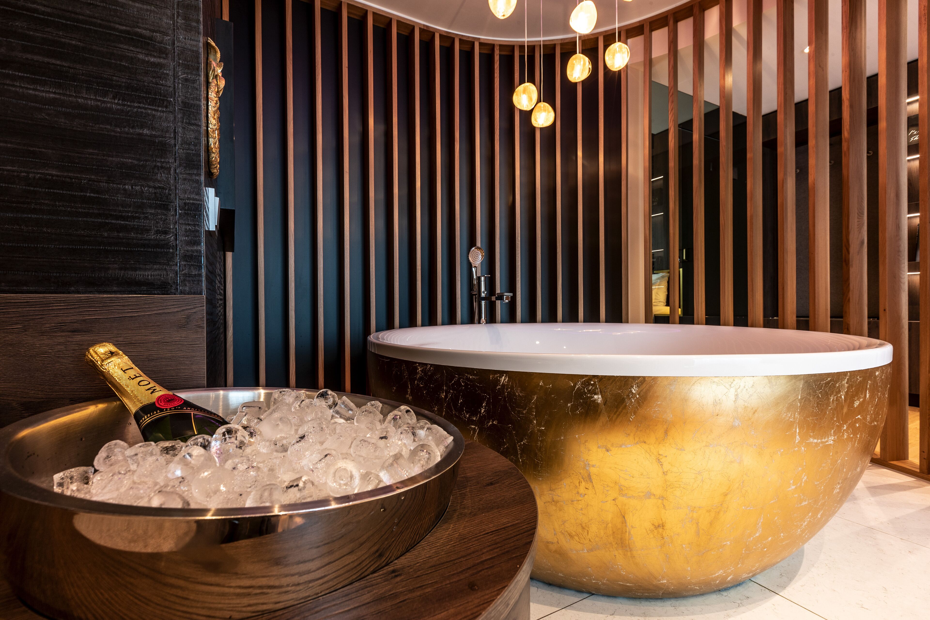 A modern bathroom with a large gold bathtub, a bowl of ice and champagne, and a wooden panel wall with vertical slats.