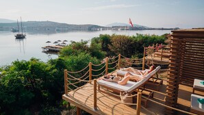 Una terraza de madera con sillones de descanso con vista a un cuerpo de agua tranquilo, una bandera y colinas distantes.