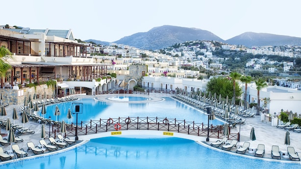 Piscina de un complejo turístico con tumbonas y sombrillas, rodeada de edificios y montañas.