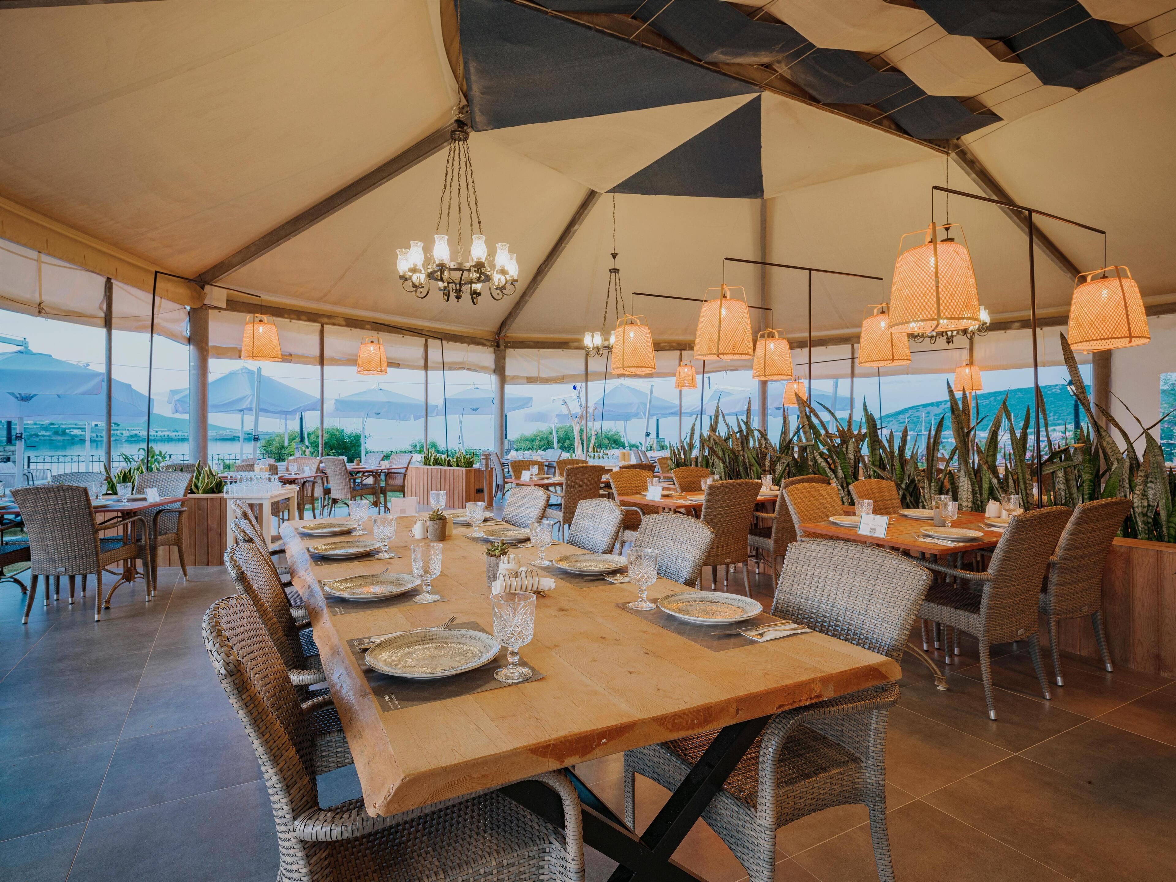 Un espace de restauration en plein air, avec des tables dressées pour les repas, entouré de chaises en osier, sous une grande tente d’où l’on peut admirer la vue sur les montagnes.