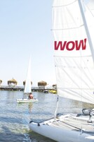Un velero con una vela blanca grande y la palabra “WOW” en ella, atracado cerca de un muelle con construcciones de techo de paja.