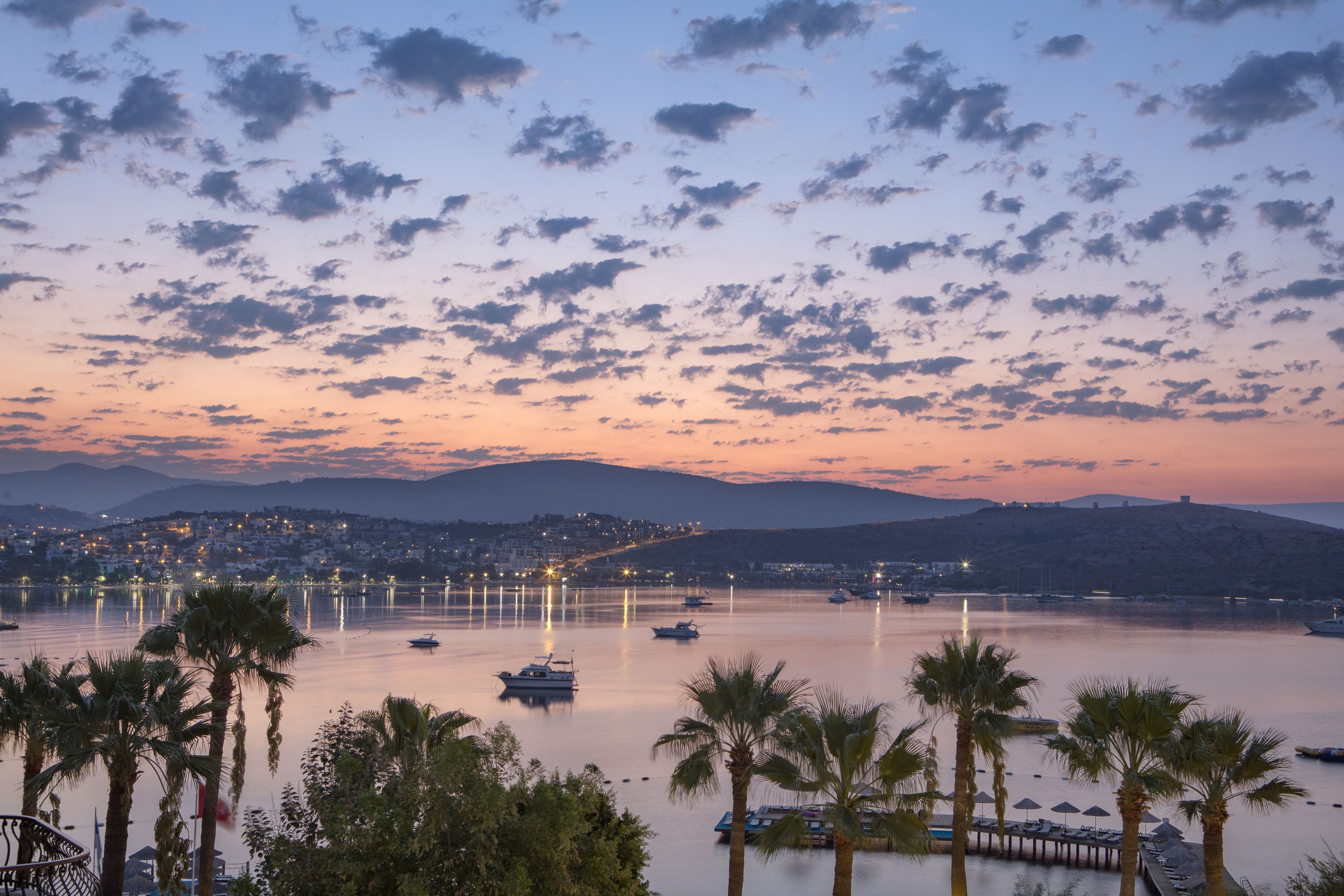 Un paysage urbain côtier au crépuscule, avec des bateaux sur des eaux calmes, des palmiers et une chaîne de montagnes lointaine.