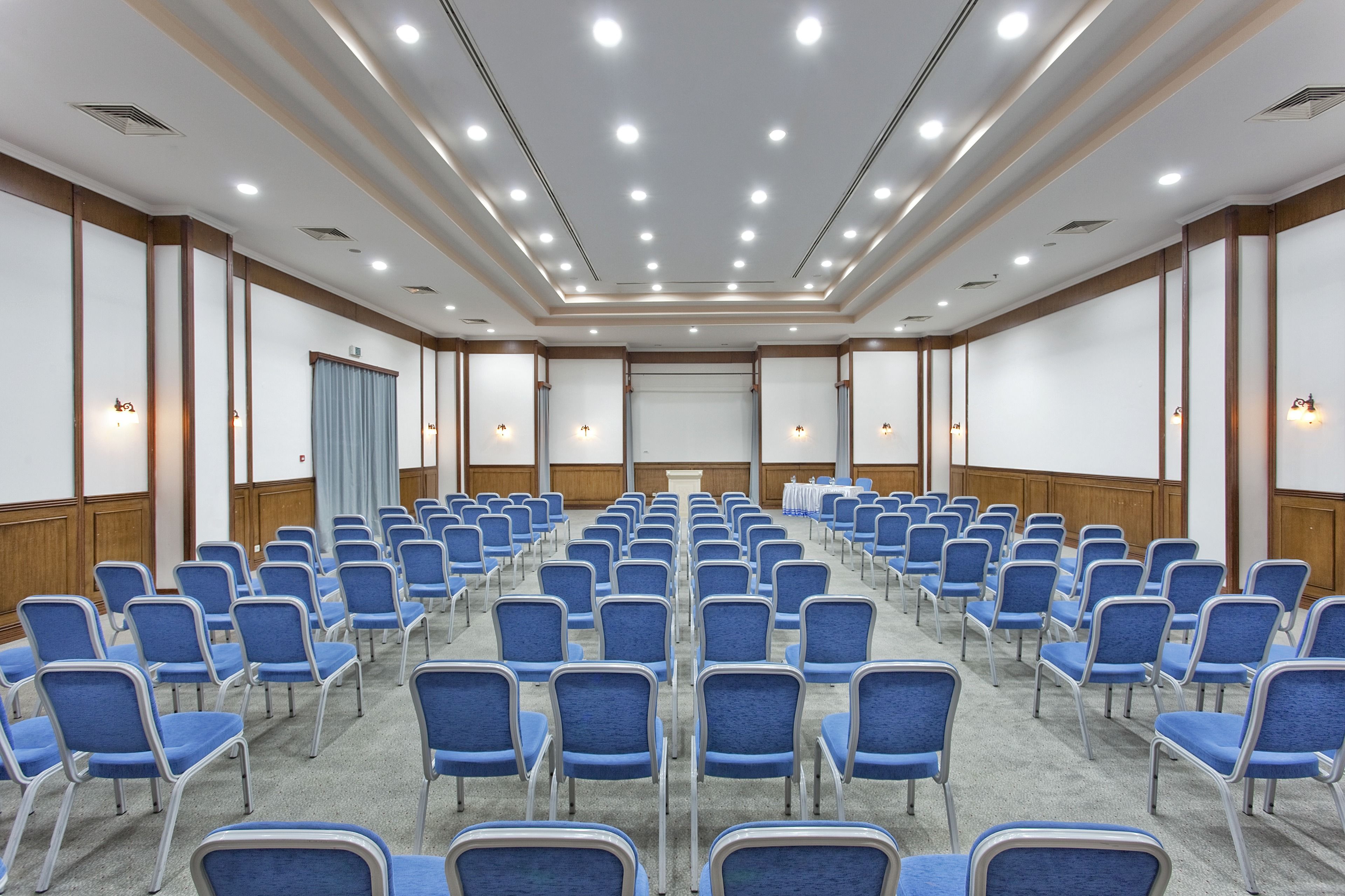 Une salle de conférence avec des rangées de chaises bleues, un pupitre et un écran à l’avant.