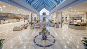 Un amplio vestíbulo de hotel con techo alto y tragaluces, una zona de recepción, áreas de descanso y un elemento decorativo central.