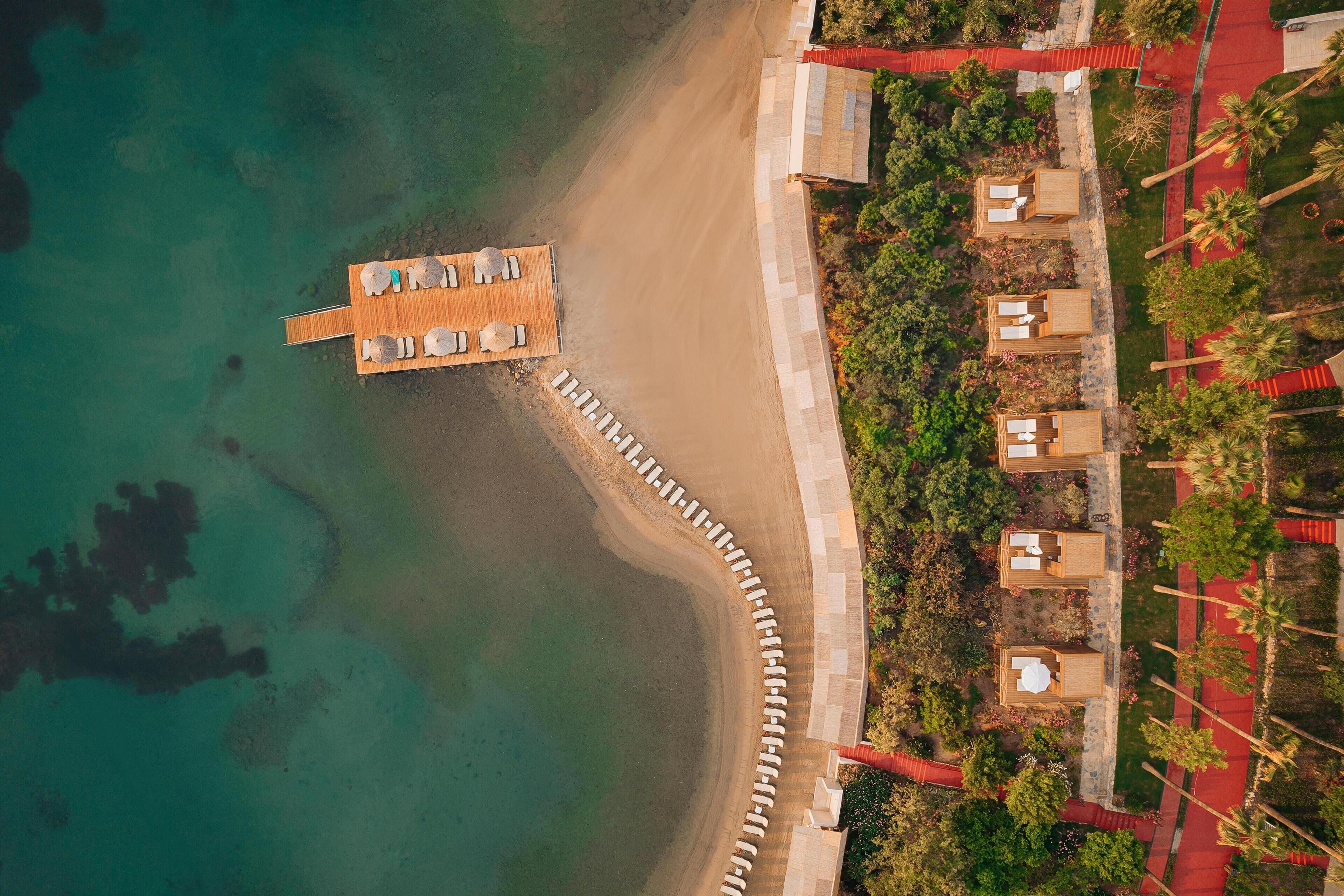 An aerial view of a beachside resort with a wooden pier leading to a deck with sun loungers, surrounded by palm trees and a red pathway.