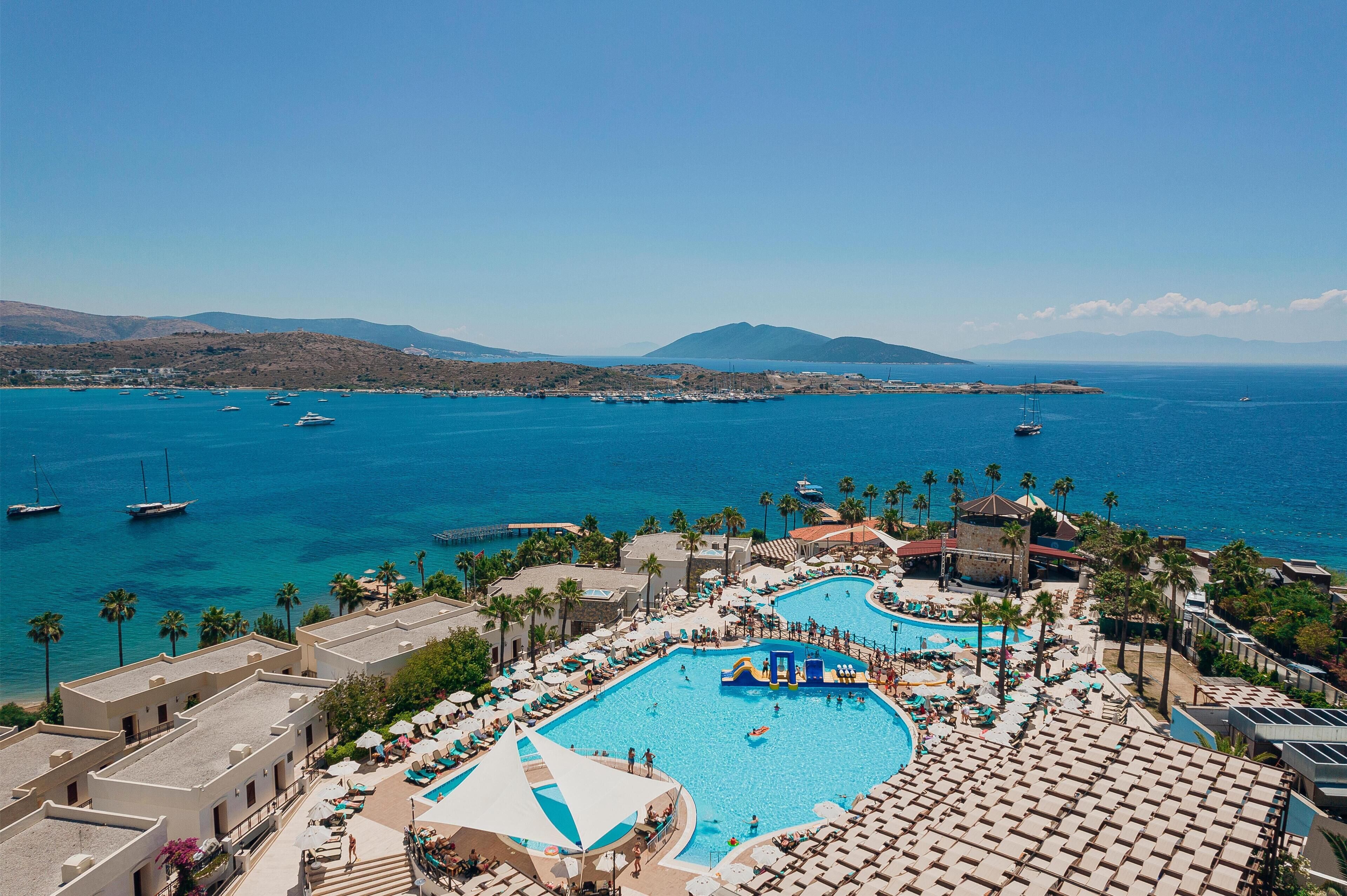 A resort with a large pool, palm trees, and boats in the background.