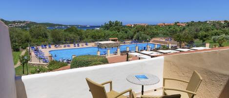 Un balcon doté de fauteuils en osier et d’une petite table, donnant sur un espace piscine avec des chaises longues et une vue sur la mer.