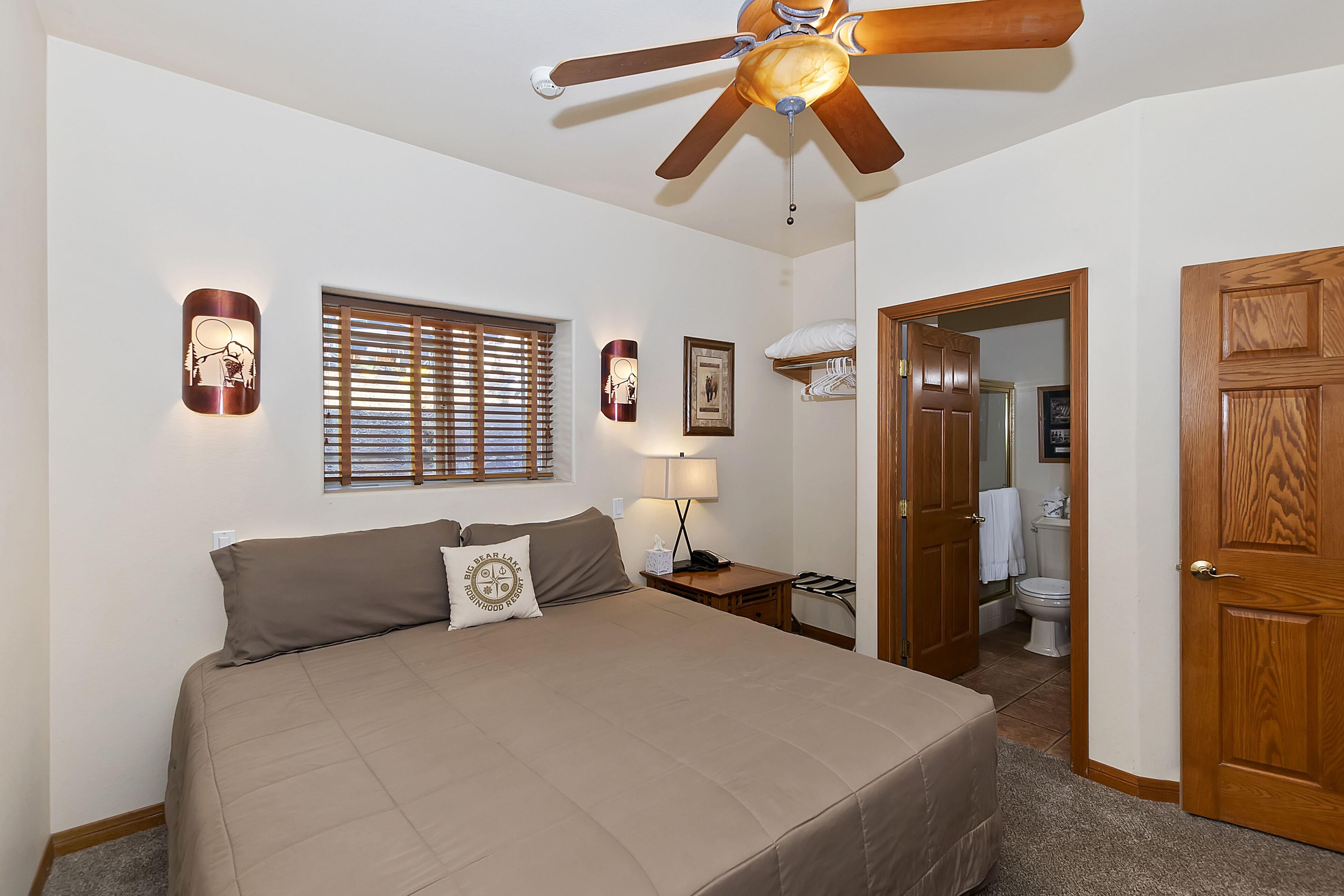 A bedroom with a large bed, a ceiling fan, a bedside table with a lamp, and a window with blinds.