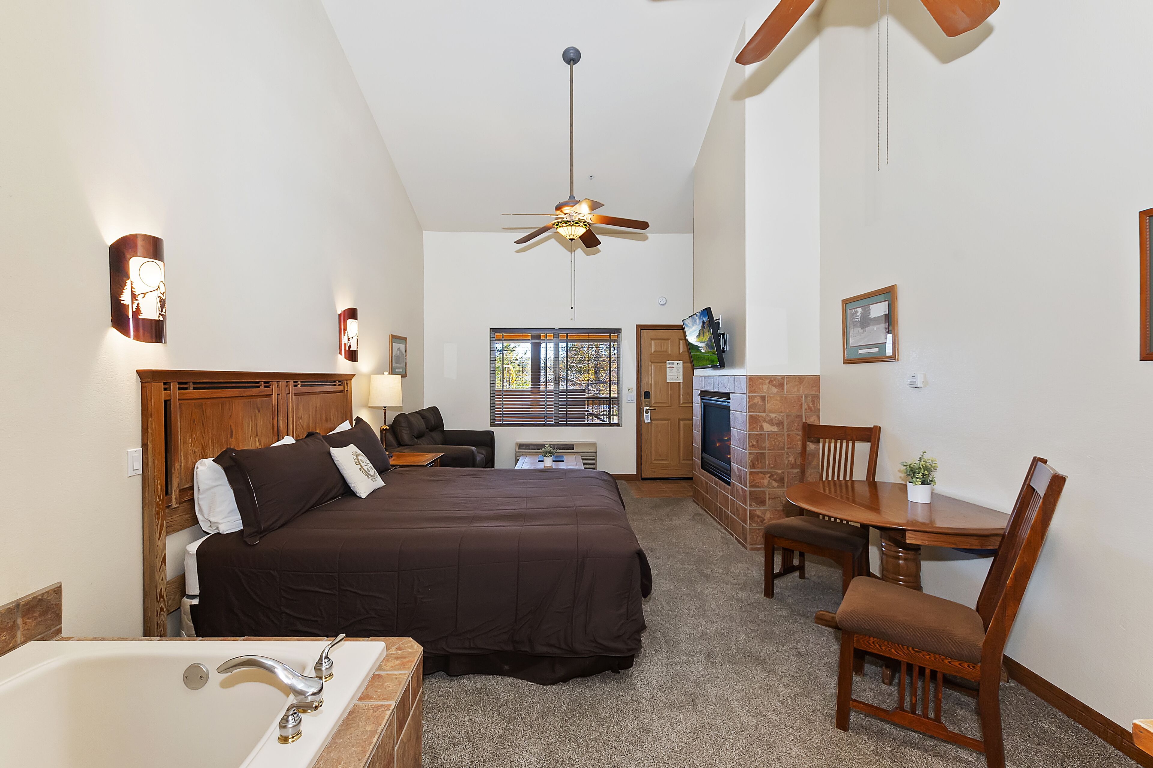 A hotel room with a large bed, a desk, a chair, a TV, and a ceiling fan.