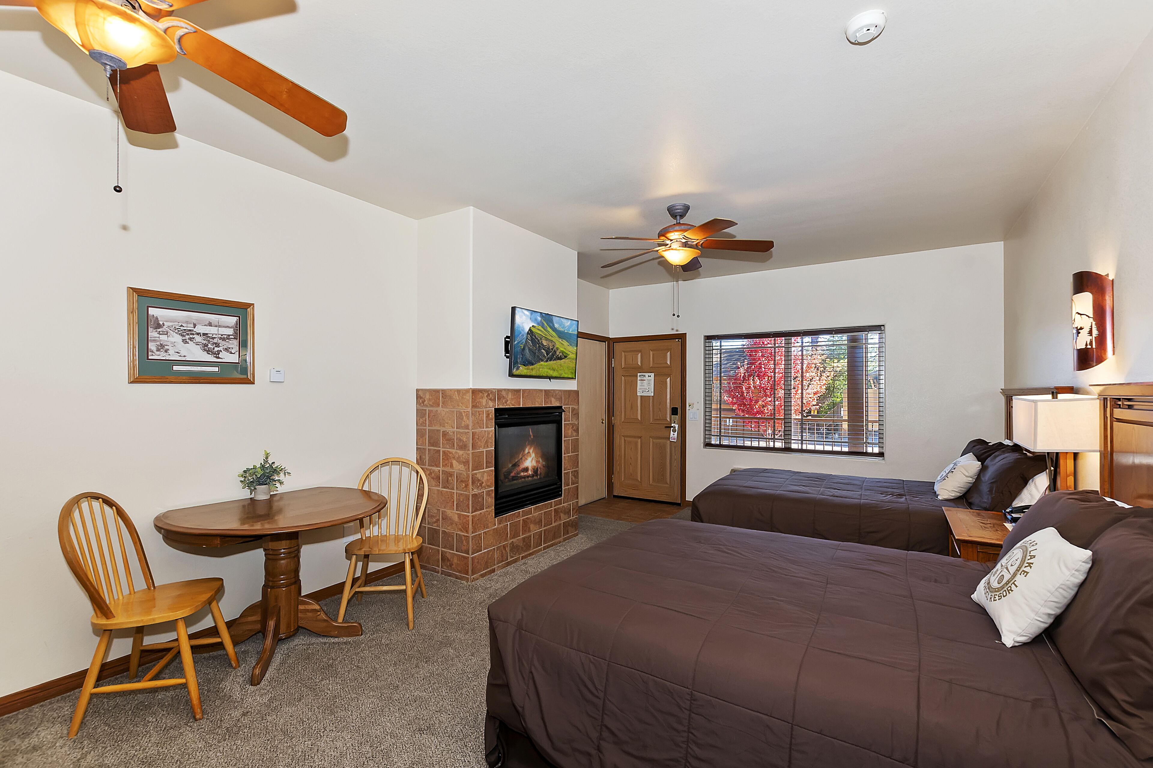 A hotel room with a fireplace, two beds, a dining table with chairs, and a ceiling fan.