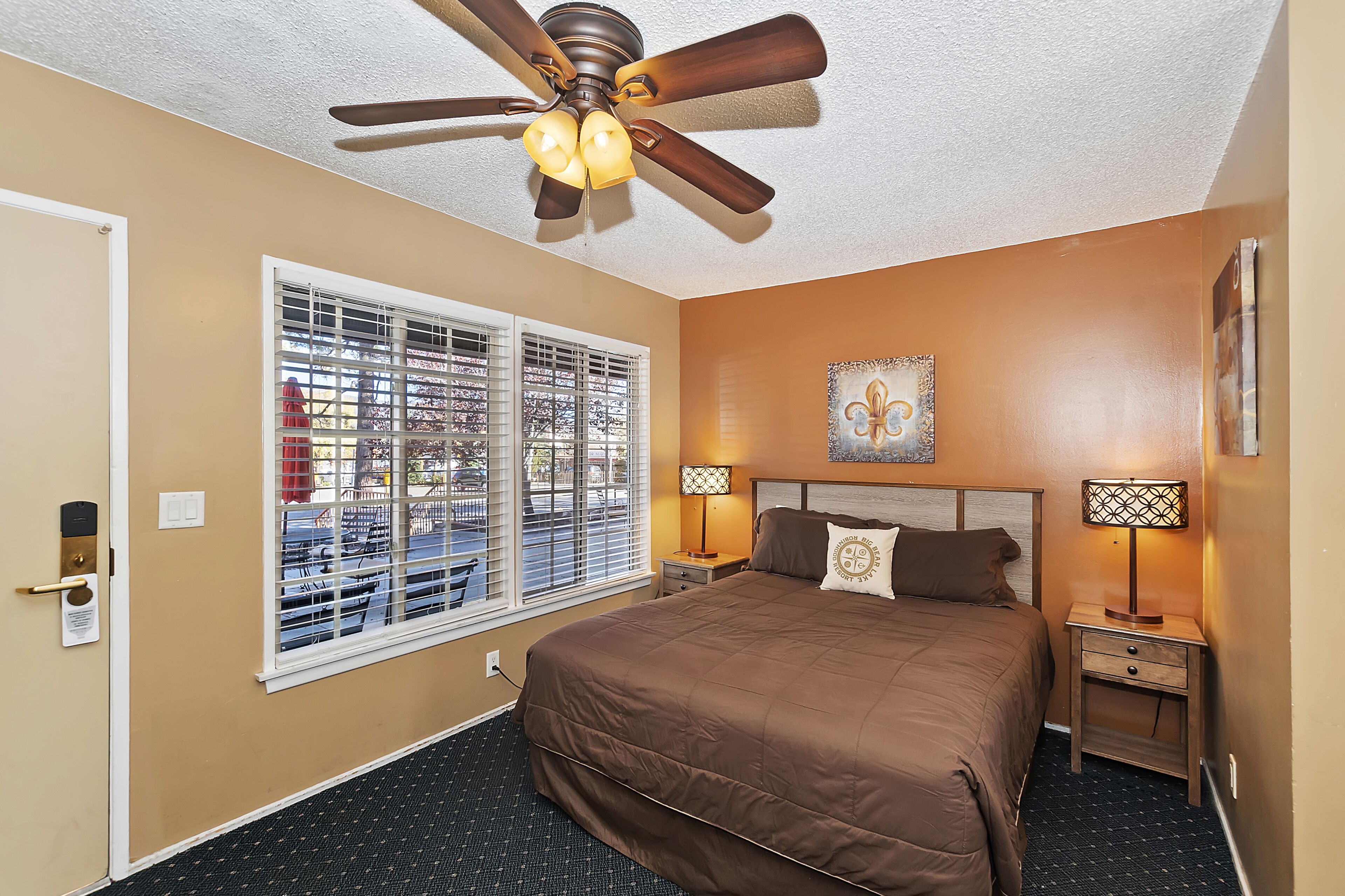 A hotel room with a large bed, two bedside tables with lamps, a ceiling fan, a window with blinds, and a painting on the wall.