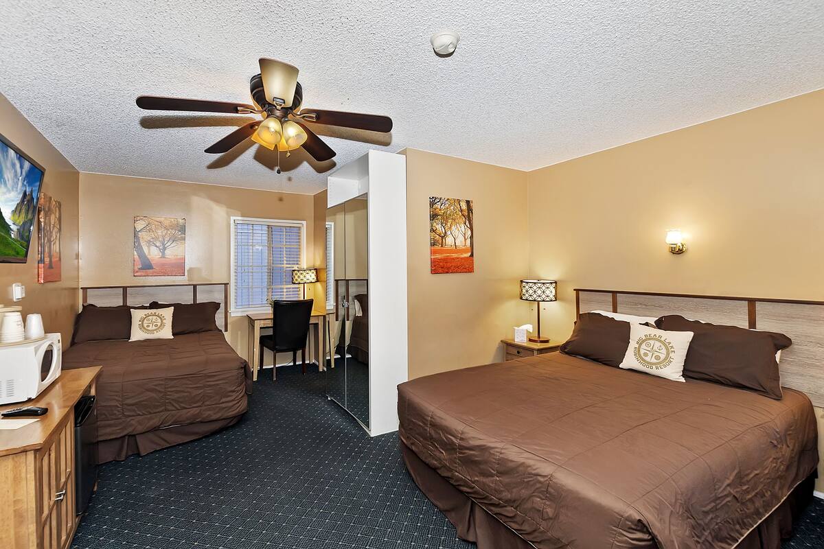 Une chambre d’hôtel avec deux lits, un bureau, une chaise, un ventilateur de plafond et des œuvres d’art accrochées aux murs.