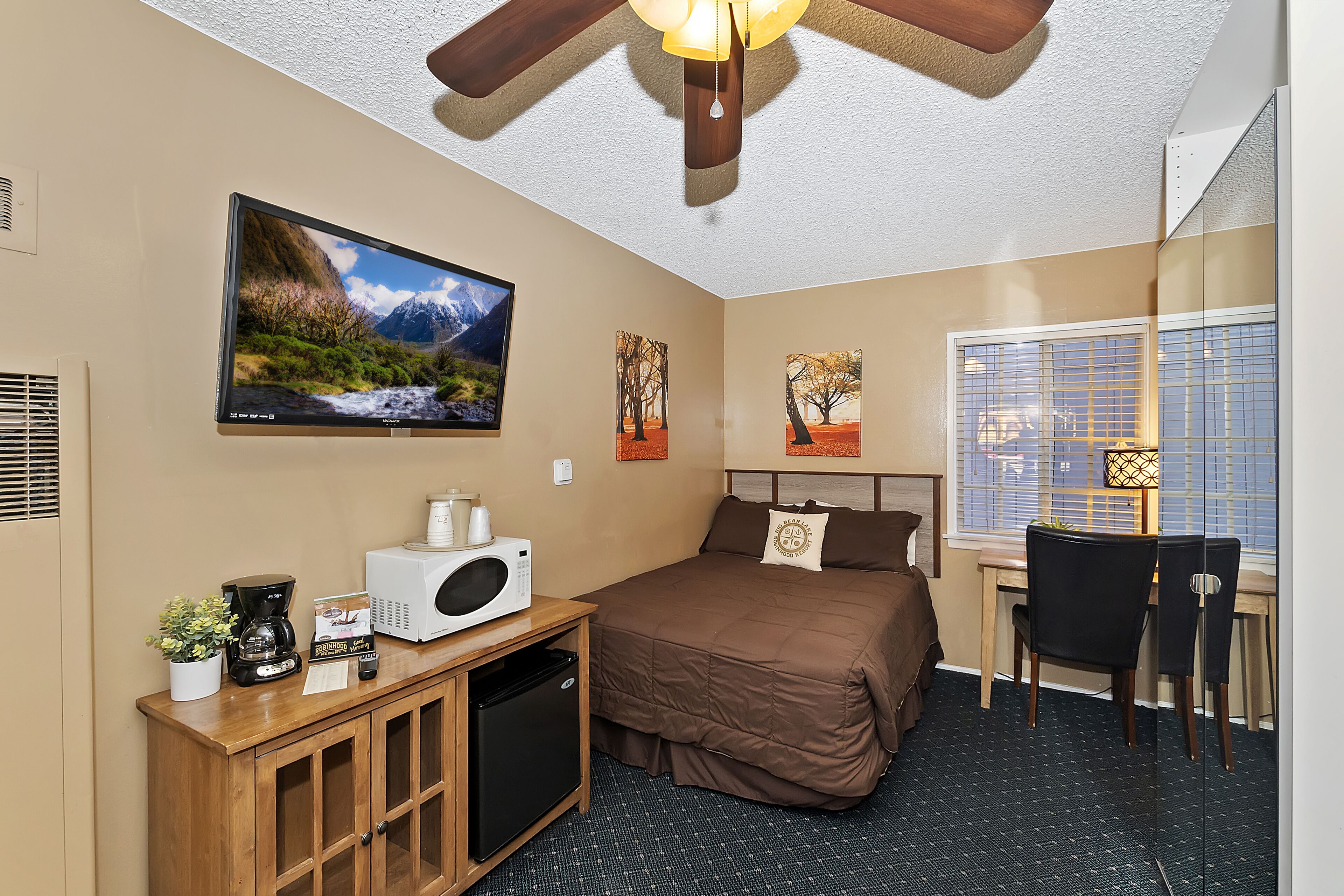 A hotel room with a bed, a desk with a chair, a television mounted on the wall, a ceiling fan, and a window with blinds.