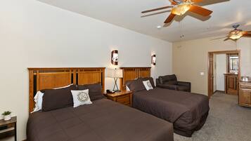 A hotel room with two beds, wooden headboards, a ceiling fan, a desk with a lamp, and a small kitchenette.