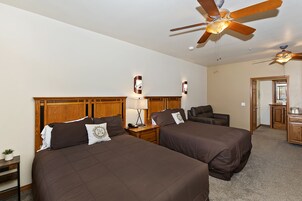 A hotel room with two beds, wooden headboards, a ceiling fan, a desk with a lamp, and a small kitchenette.