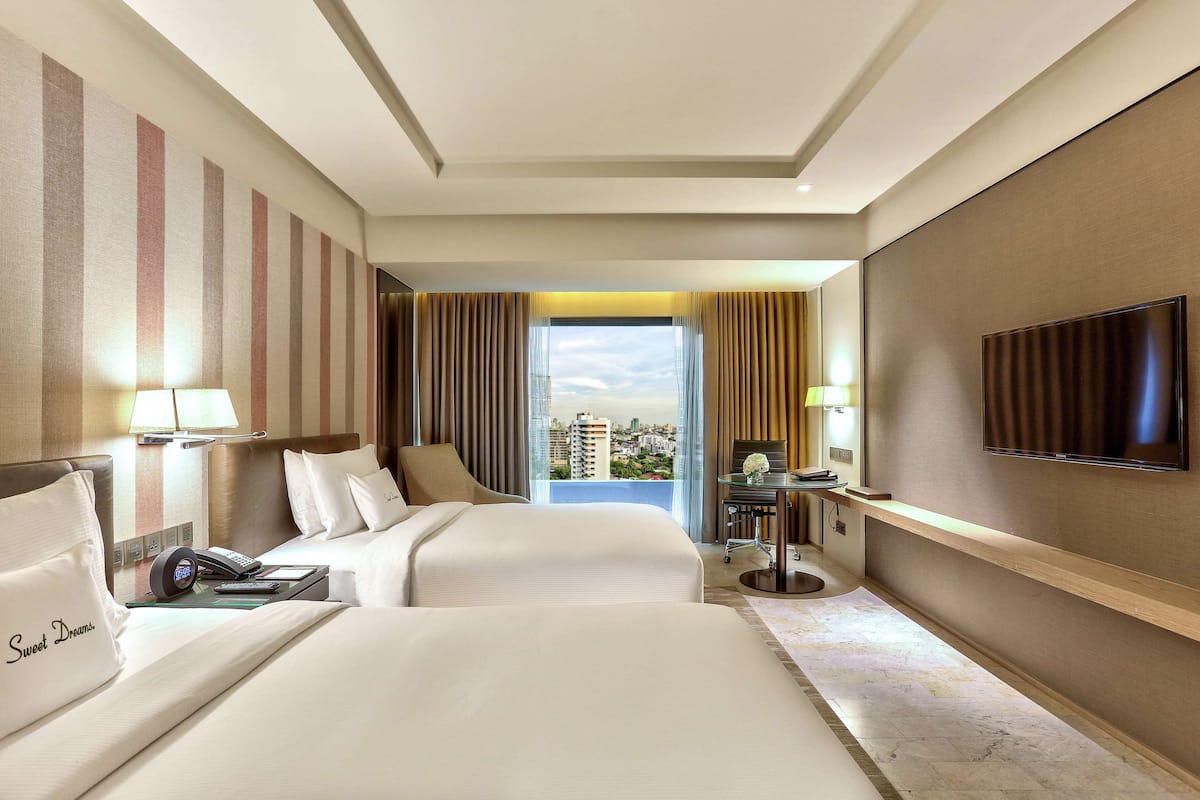 A hotel room with two beds, a TV, a desk with a chair, and a view of the city.