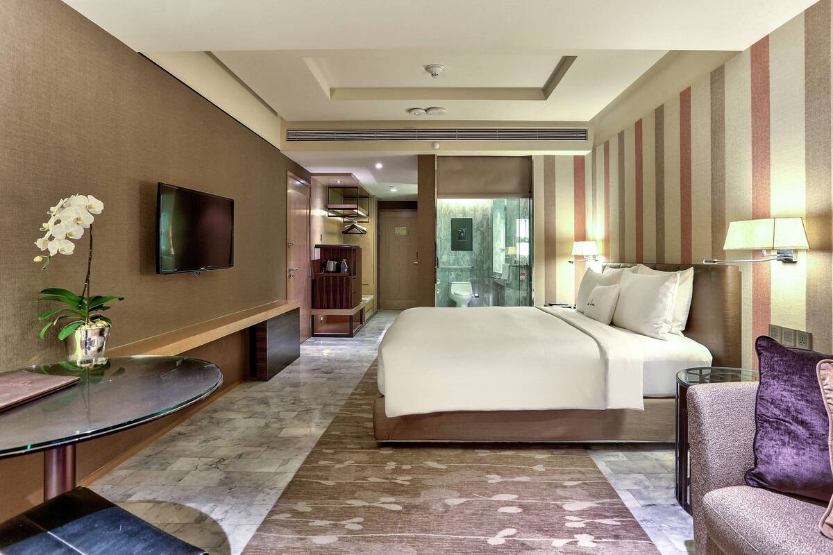 A hotel room with a bed, a TV, a desk with a vase of flowers, a chair, and a bathroom visible through a glass door.