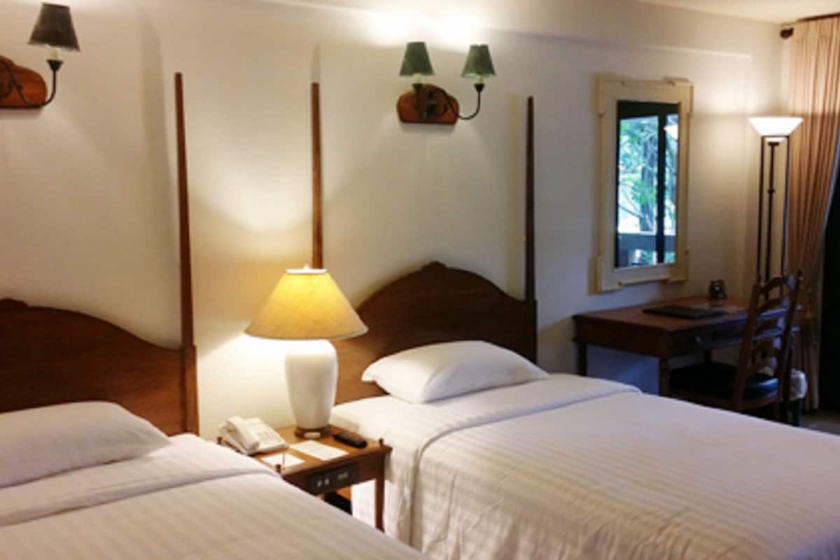 Une chambre d’hôtel avec deux lits simples, une table de chevet avec une lampe, un bureau avec une chaise et une fenêtre avec des rideaux.