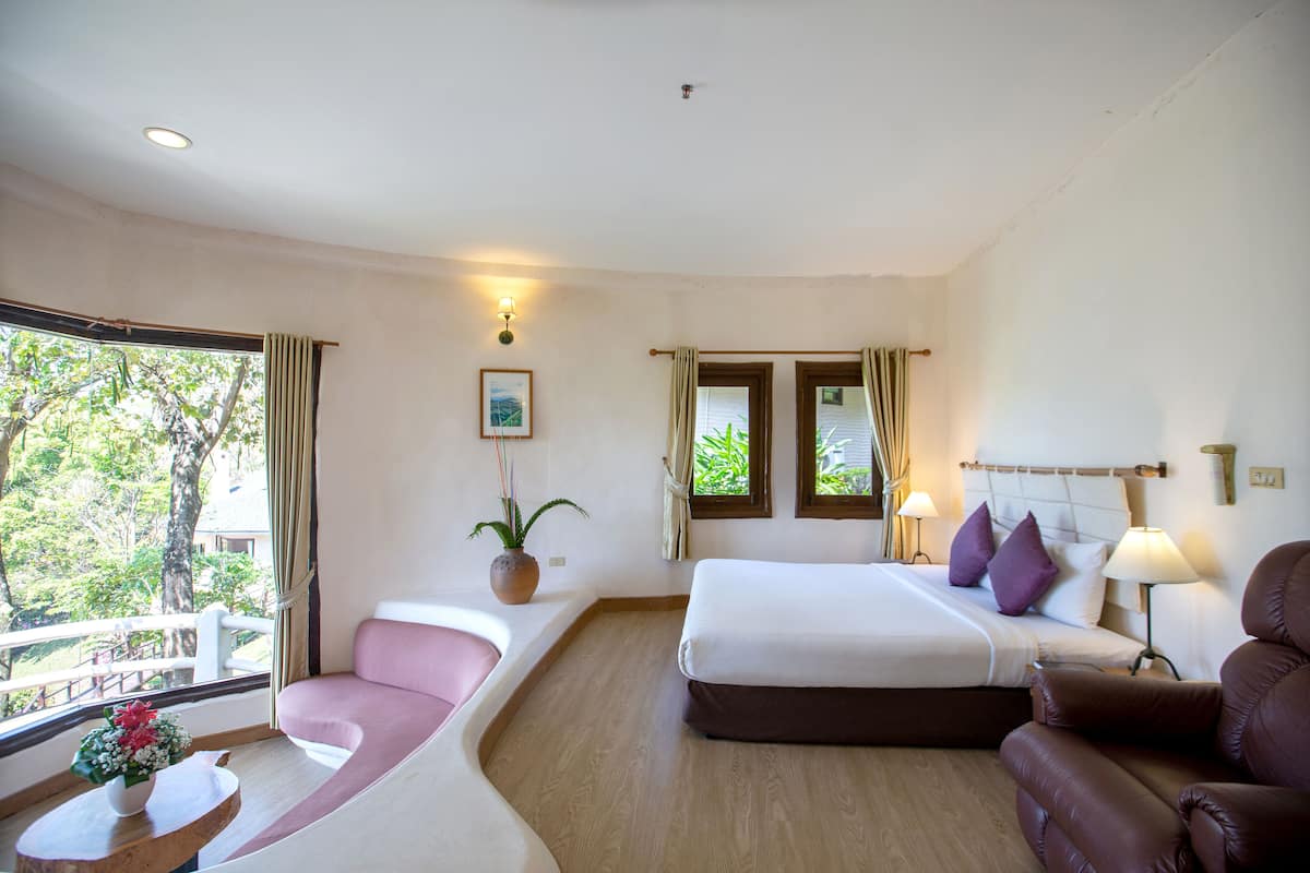 A hotel room with a bed, a sofa, a chair, a window with curtains, a bedside table with a vase, and a view of trees outside.