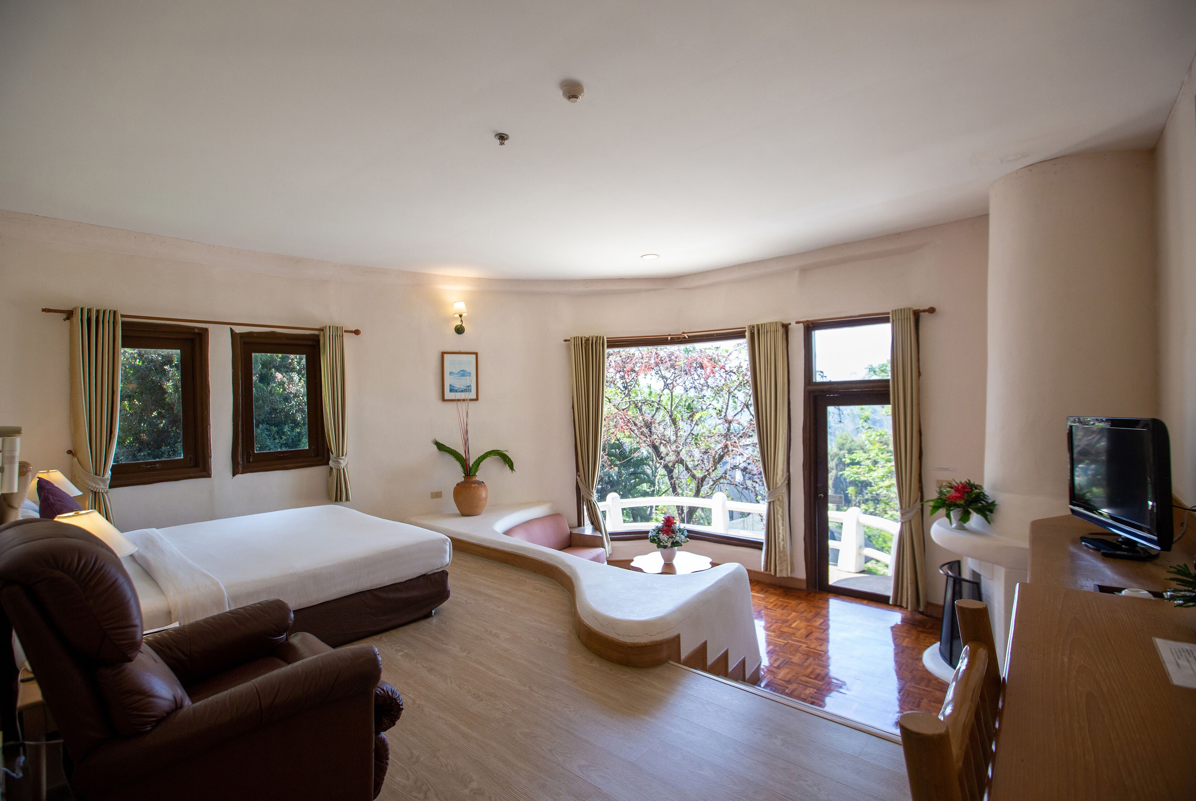 A hotel room with a bed, a sofa, a TV, and a balcony with a view of trees.
