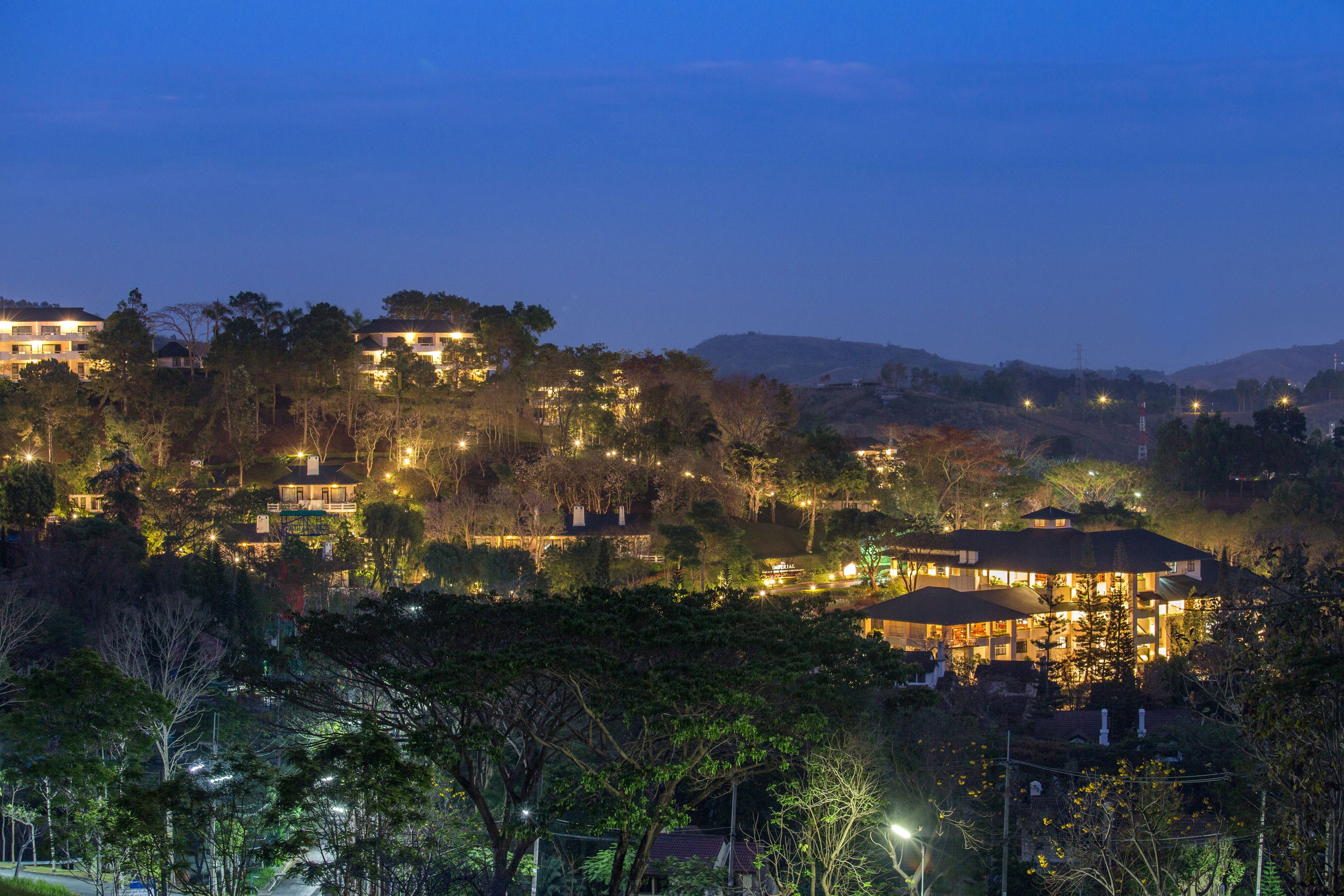 Un barrio residencial en una ladera con varias casas, cada una con ventanas iluminadas e iluminación exterior, rodeado de árboles.