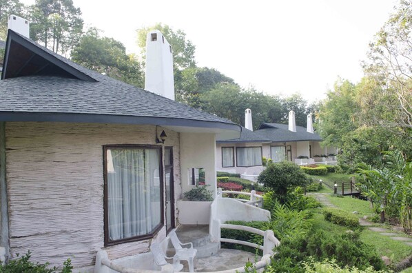 A row of cottages with a stone facade, a small porch with chairs, and a garden with a wooden bridge.