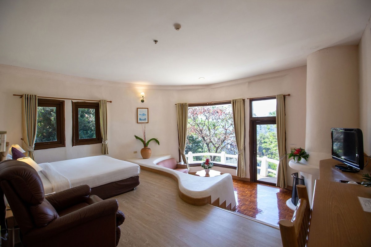A hotel room with a bed, a sofa, a TV, a small table, and a balcony with a view of trees.