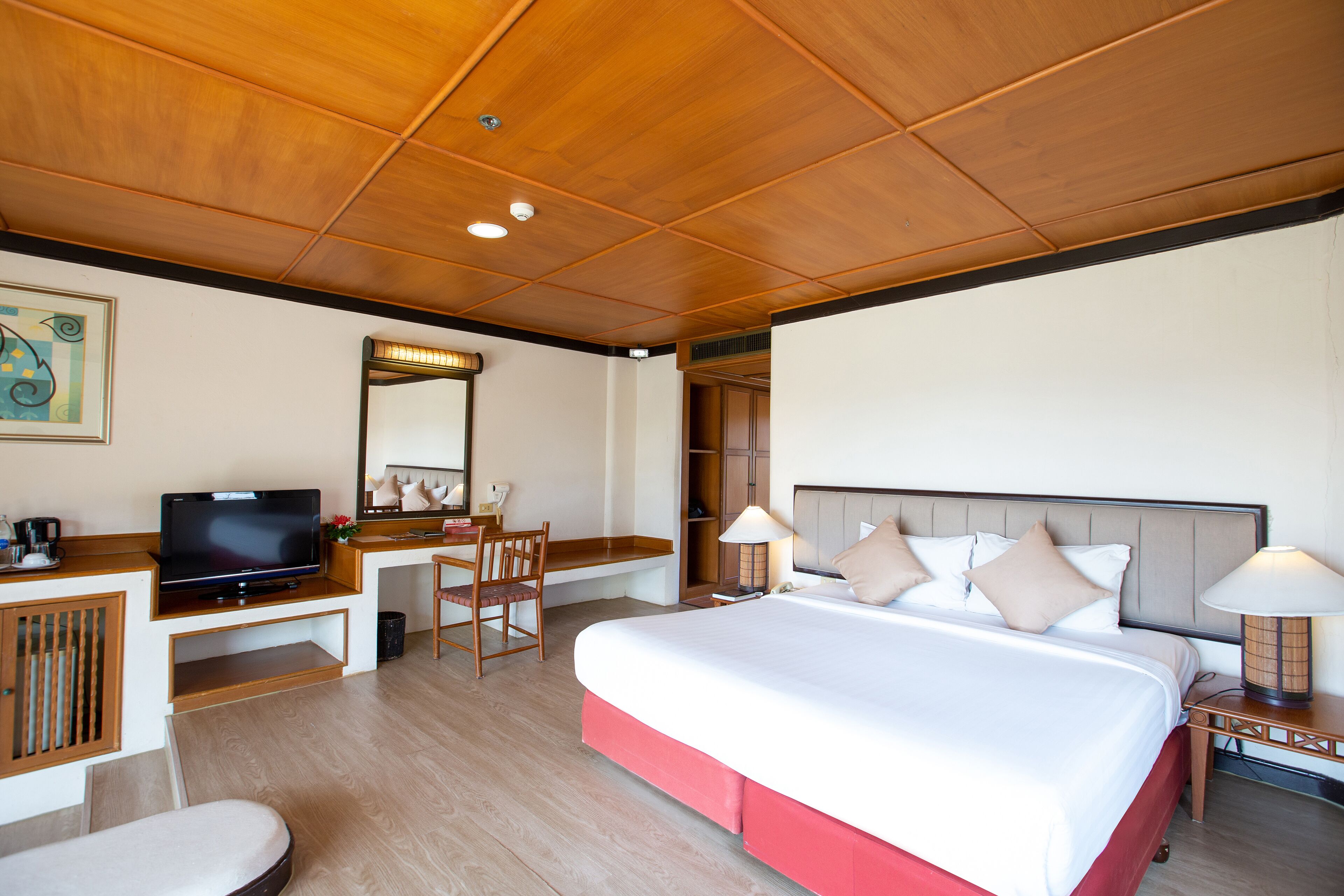Habitación de hotel con techo de madera, cama con ropa de cama blanca y roja, escritorio con televisor, una silla, una lámpara y un espejo.