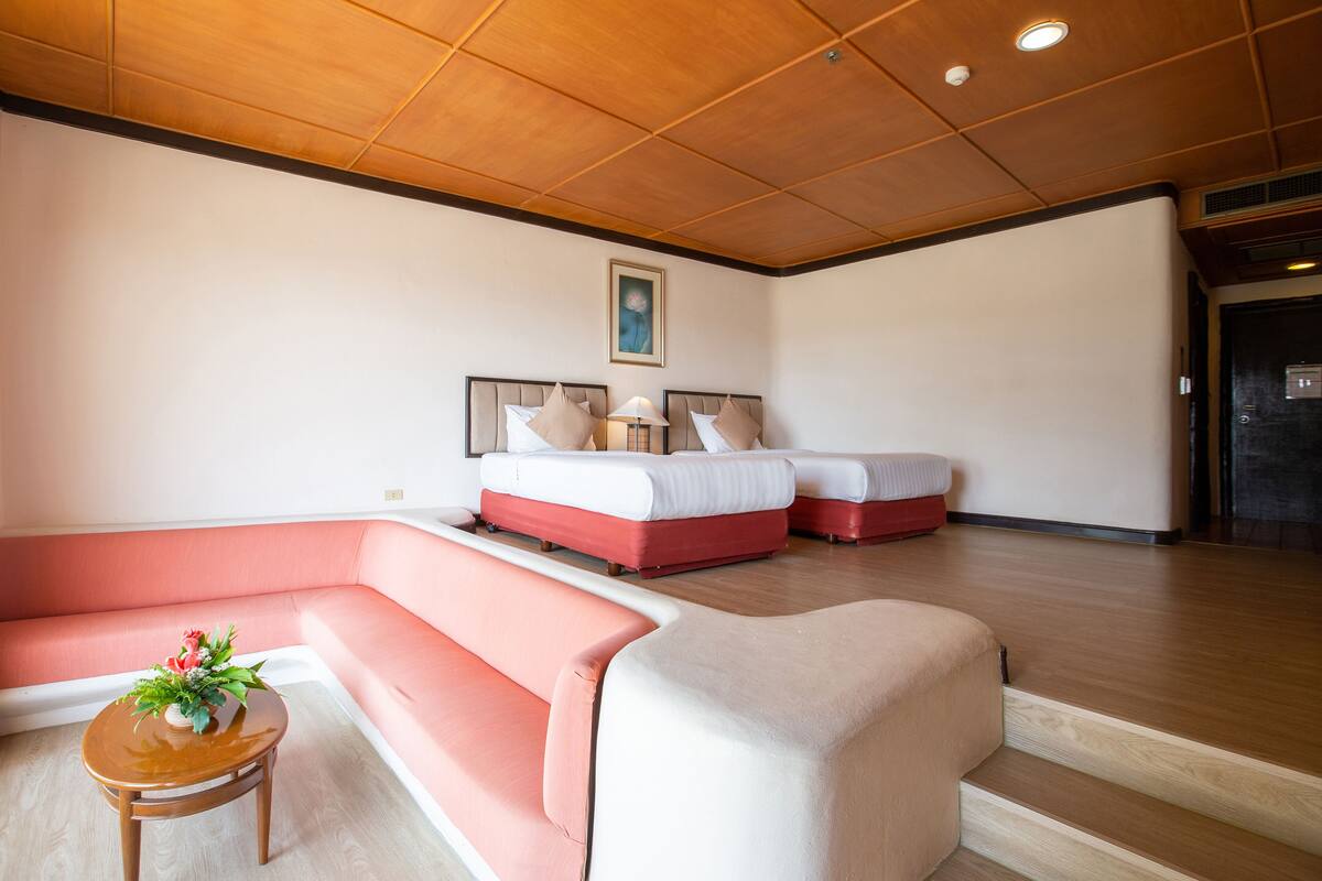 A hotel room with a wooden ceiling, two beds with red and white bedding, a pink sofa, and a small wooden table with a vase of flowers.