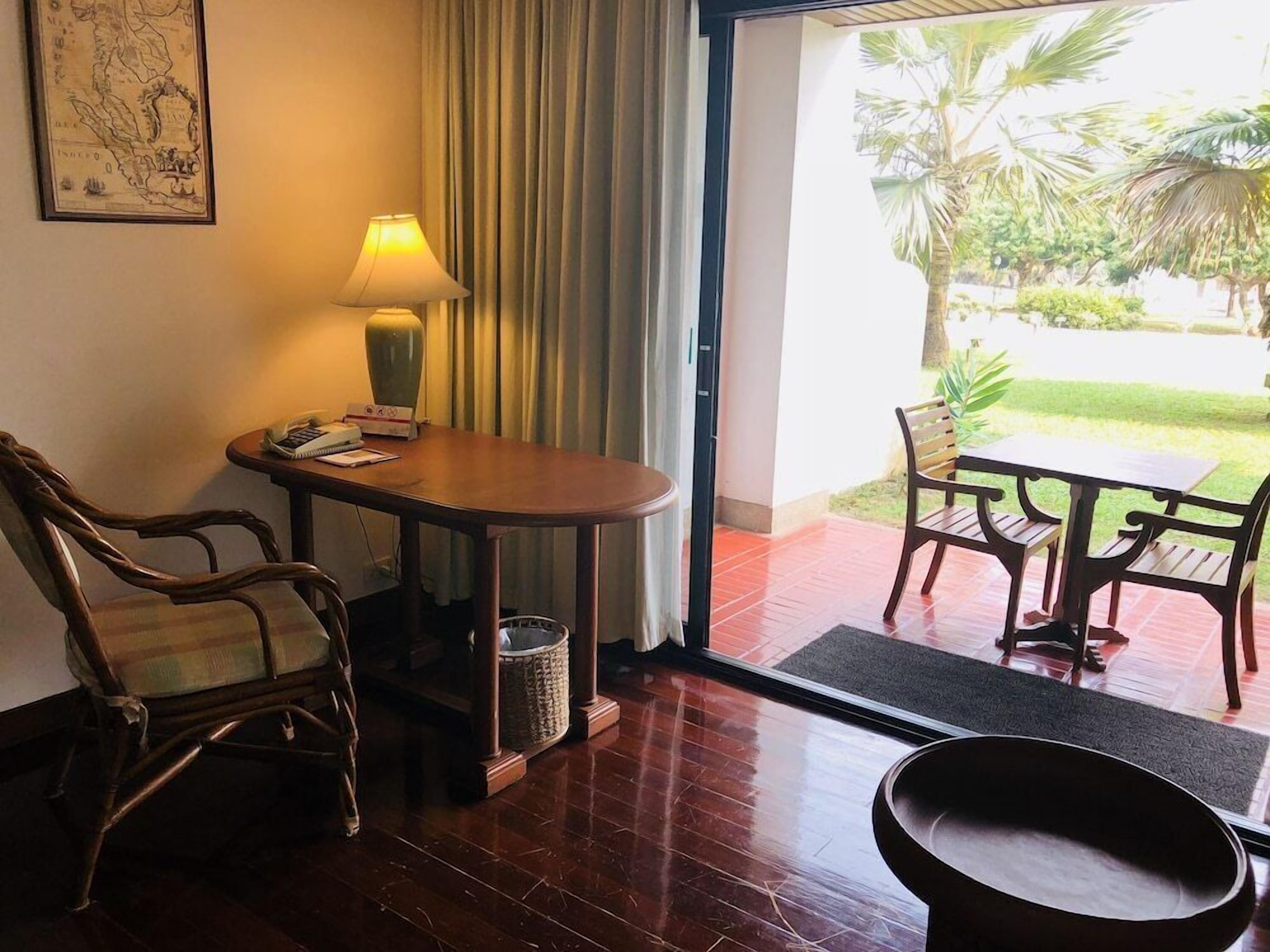 Una habitación con una mesa de madera, dos sillas, una lámpara, un mapa enmarcado y vistas a un jardín a través de un ventanal.
