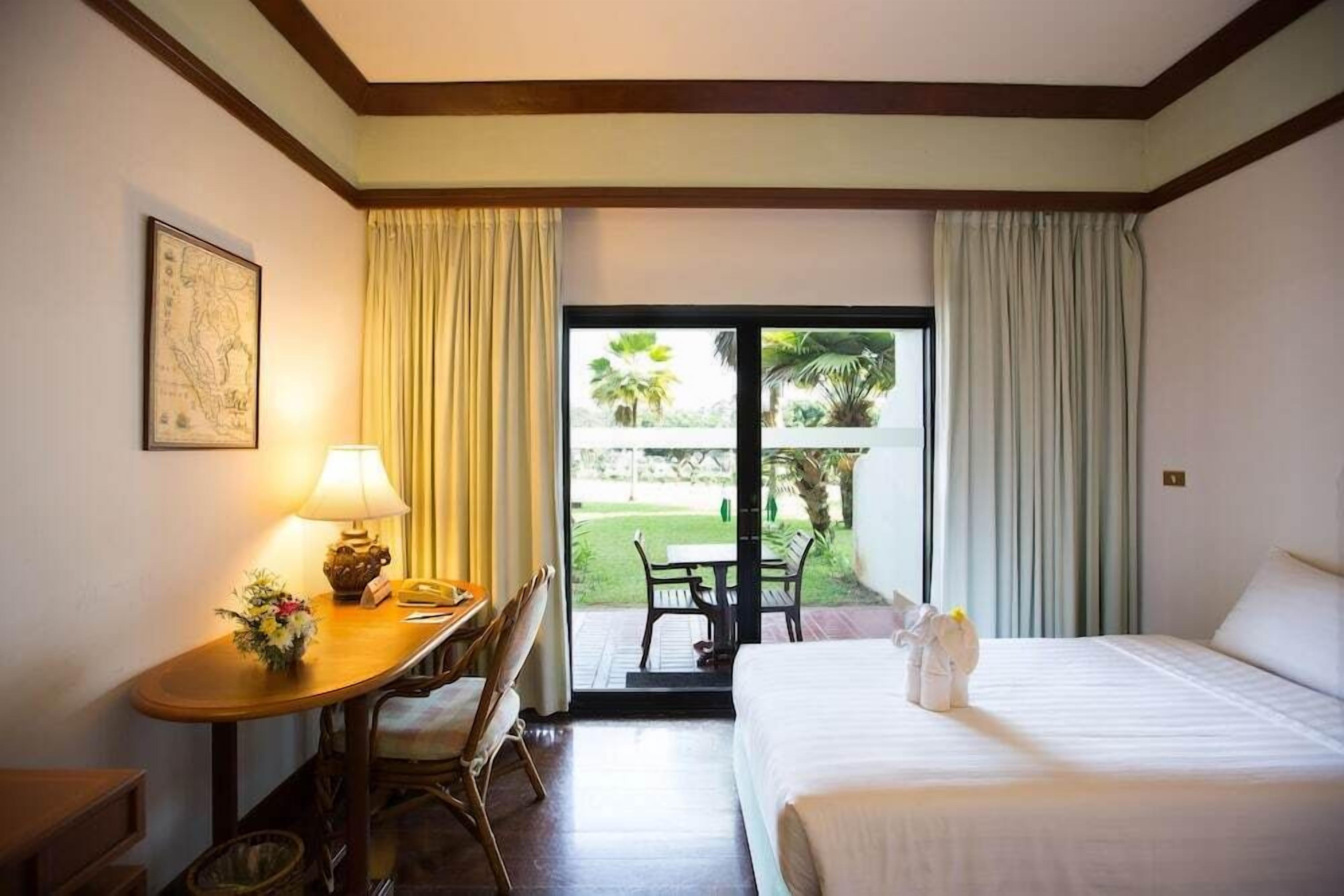 A hotel room with a bed, a desk with a lamp, a chair, a small table with a vase, and a view of the outdoors through a sliding door.
