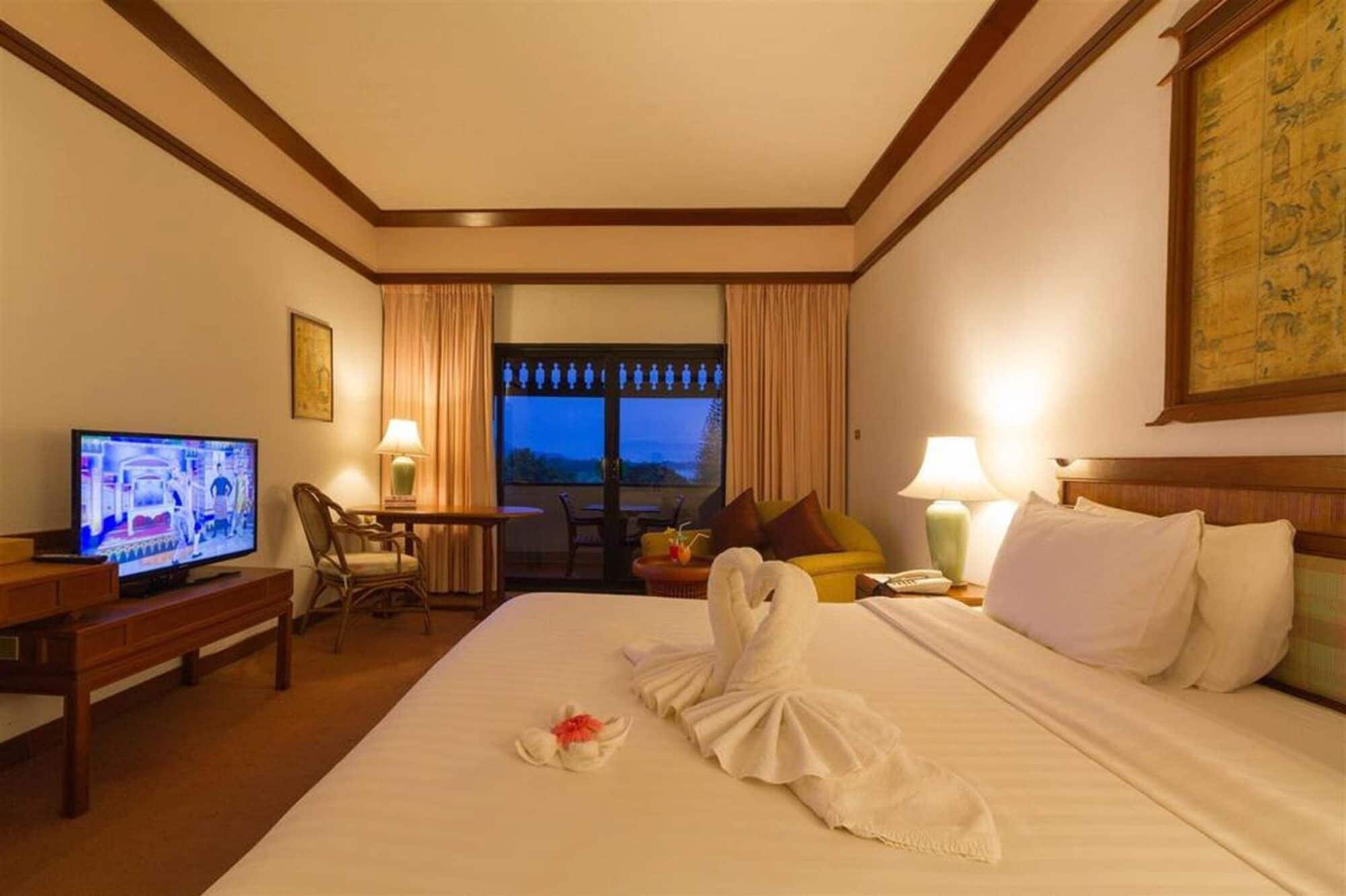 A hotel room with a bed, a TV, a desk, a chair, a window with curtains, and a painting on the wall.