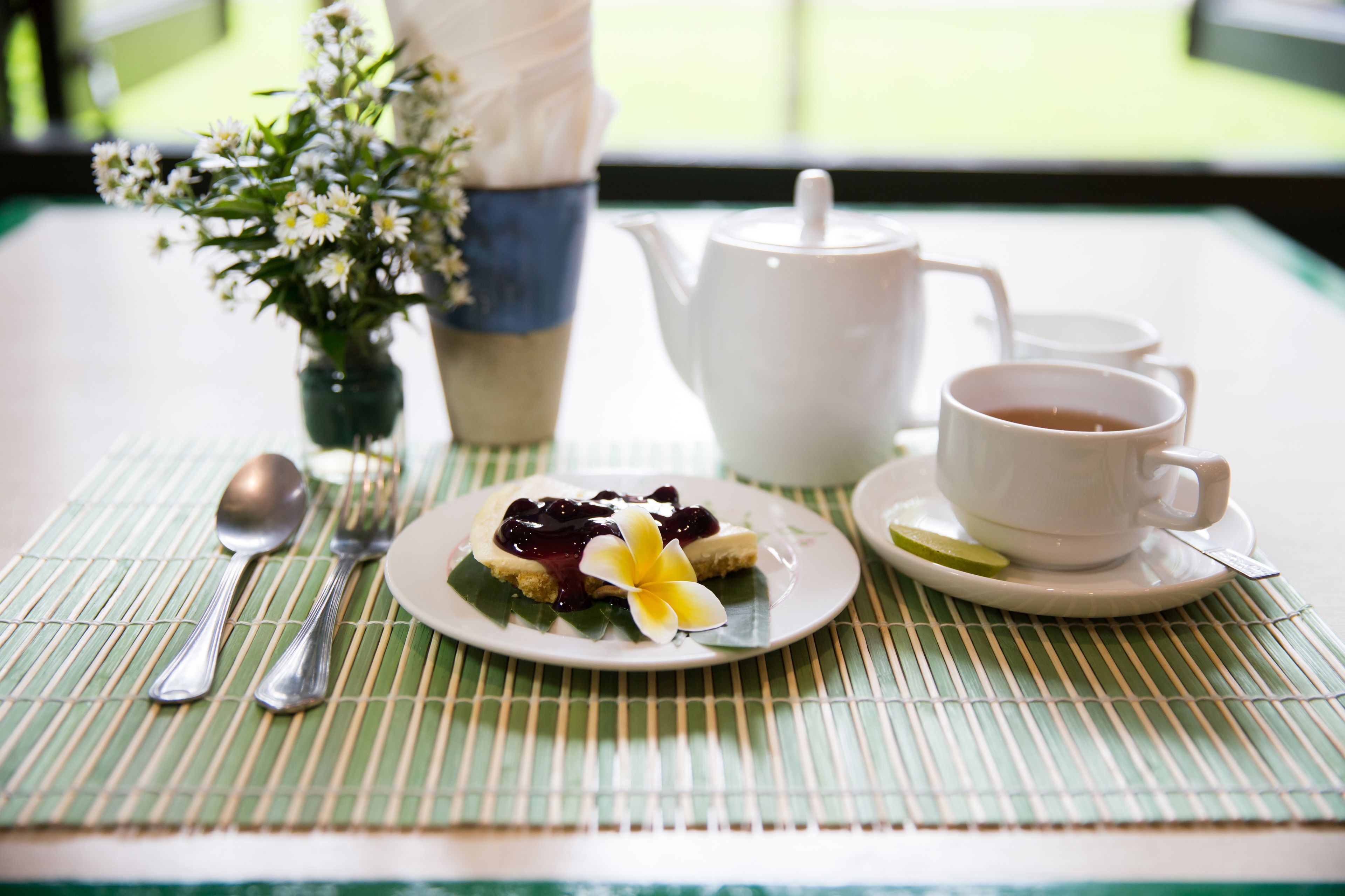 Une table avec une théière blanche, deux tasses blanches, une assiette avec un dessert, une cuillère, une fourchette et un petit vase avec des fleurs.