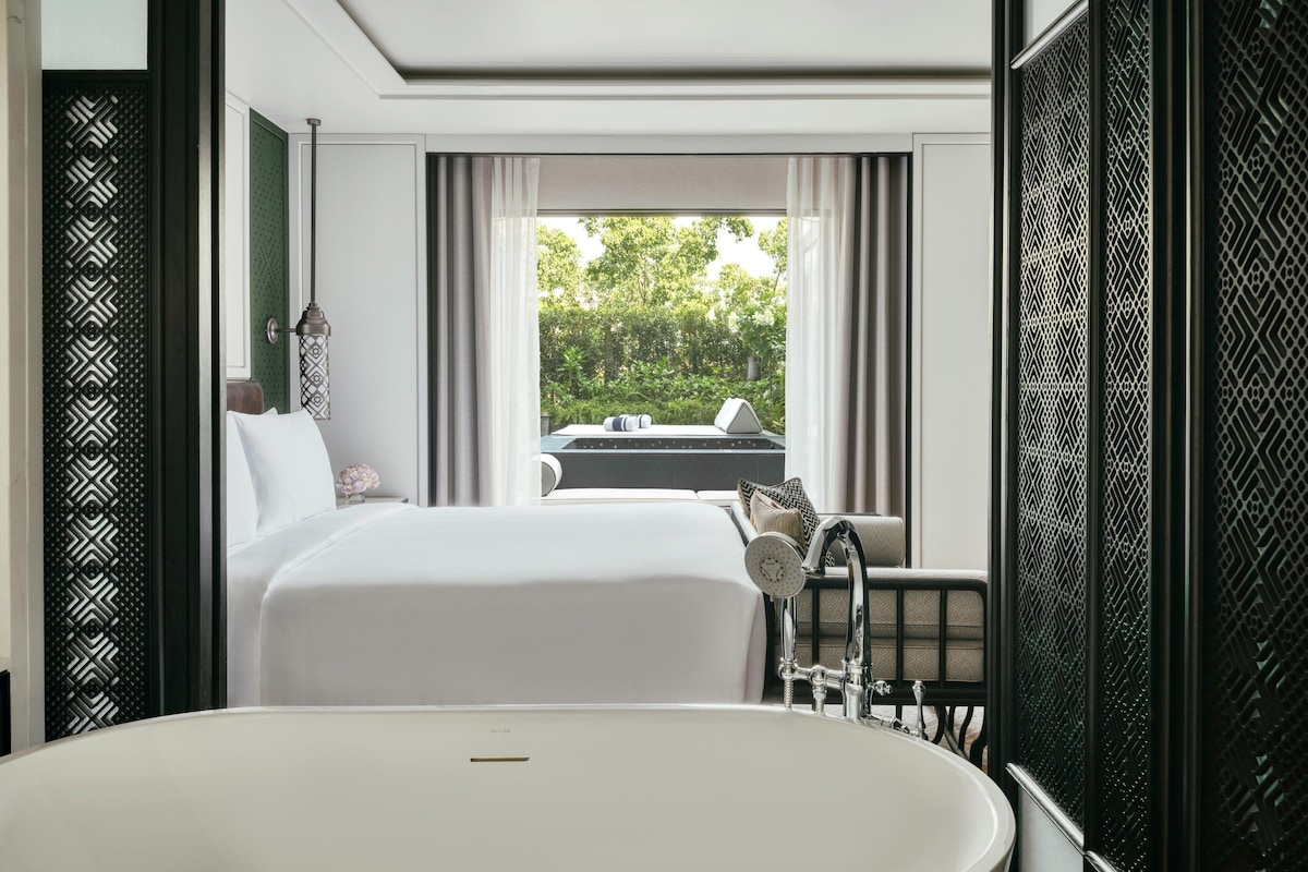 Ett modernt hotellrum med ett stort badkar, en säng med vita sängkläder och utsikt över en pool och grönområden utanför.