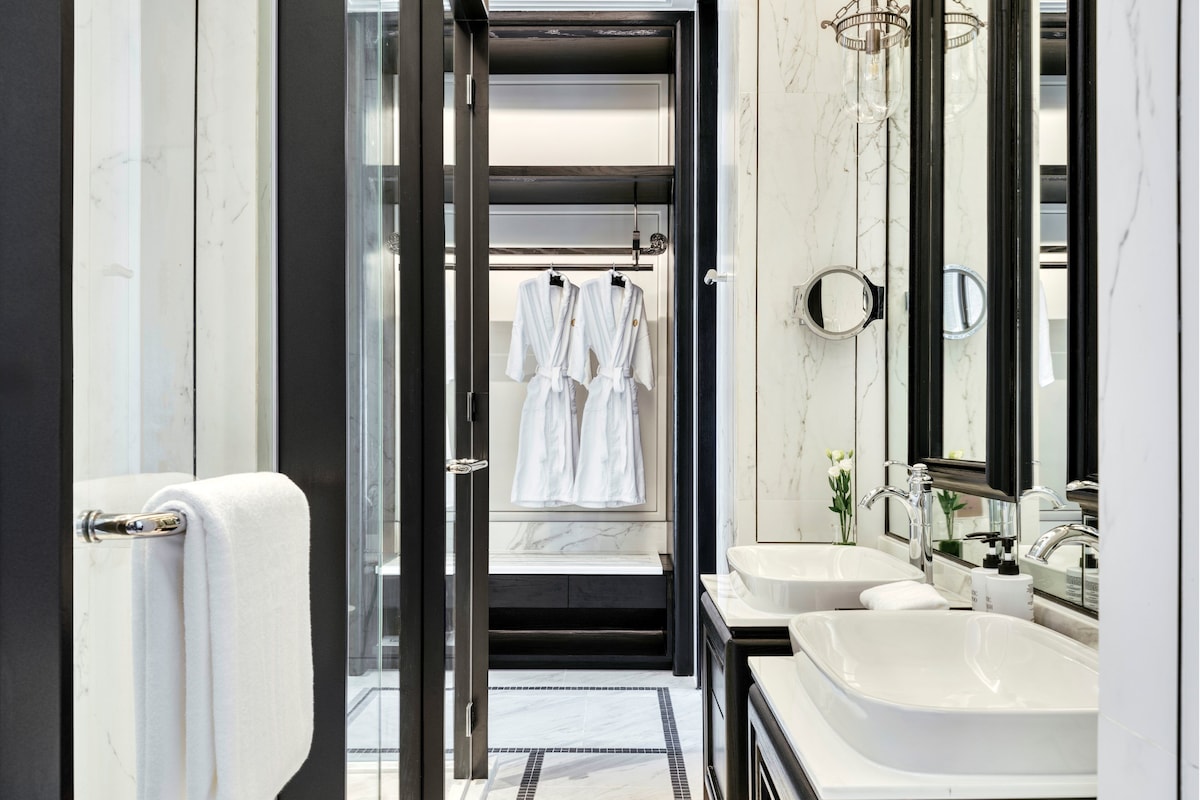 Ein modernes Badezimmer mit zwei Waschbecken, einem großen Spiegel und einem begehbaren Kleiderschrank mit aufgehängten Bademänteln.