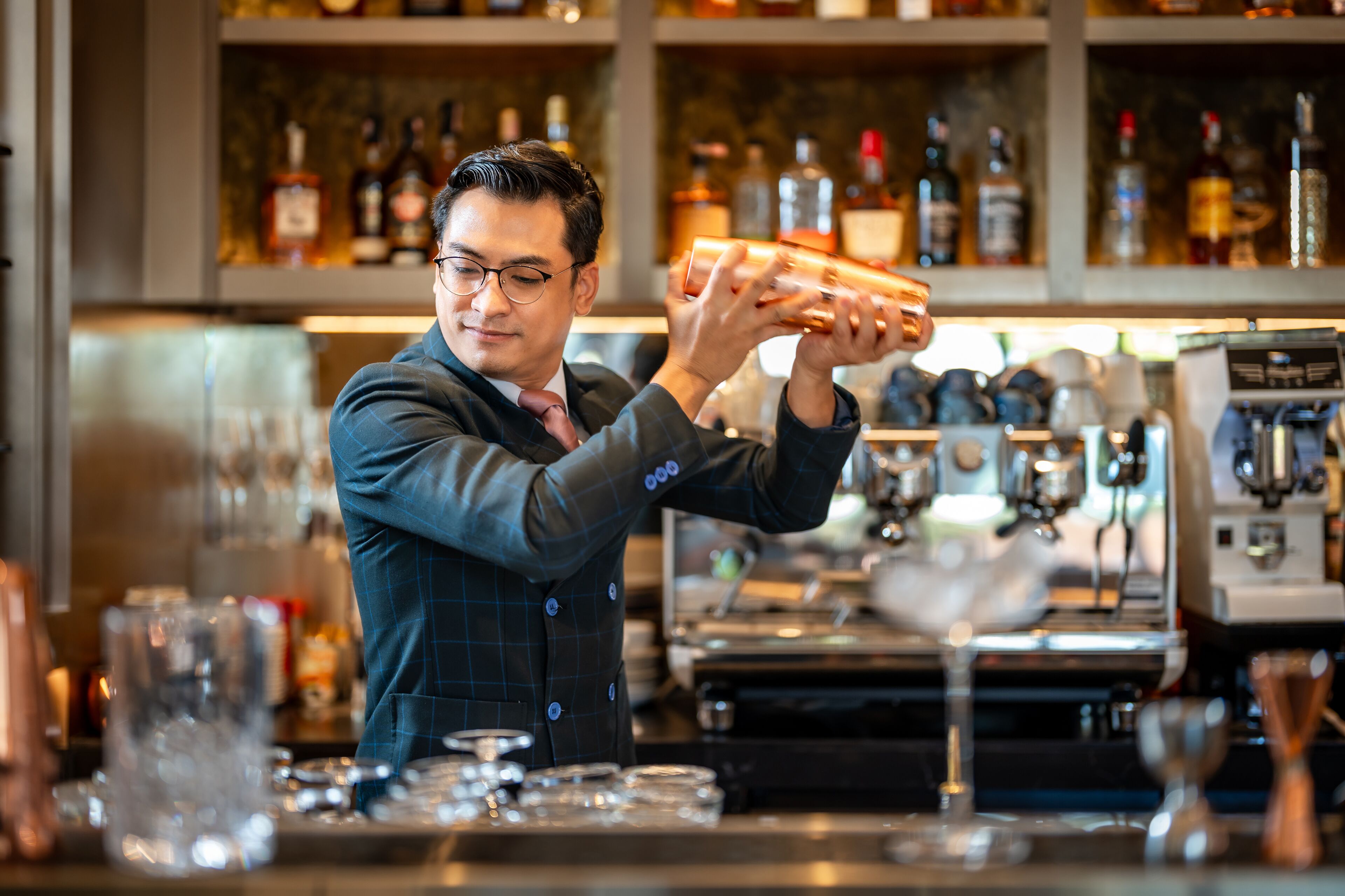 Un barista con traje oscuro prepara una bebida en la barra, con estantes llenos de botellas y máquinas de café al fondo.