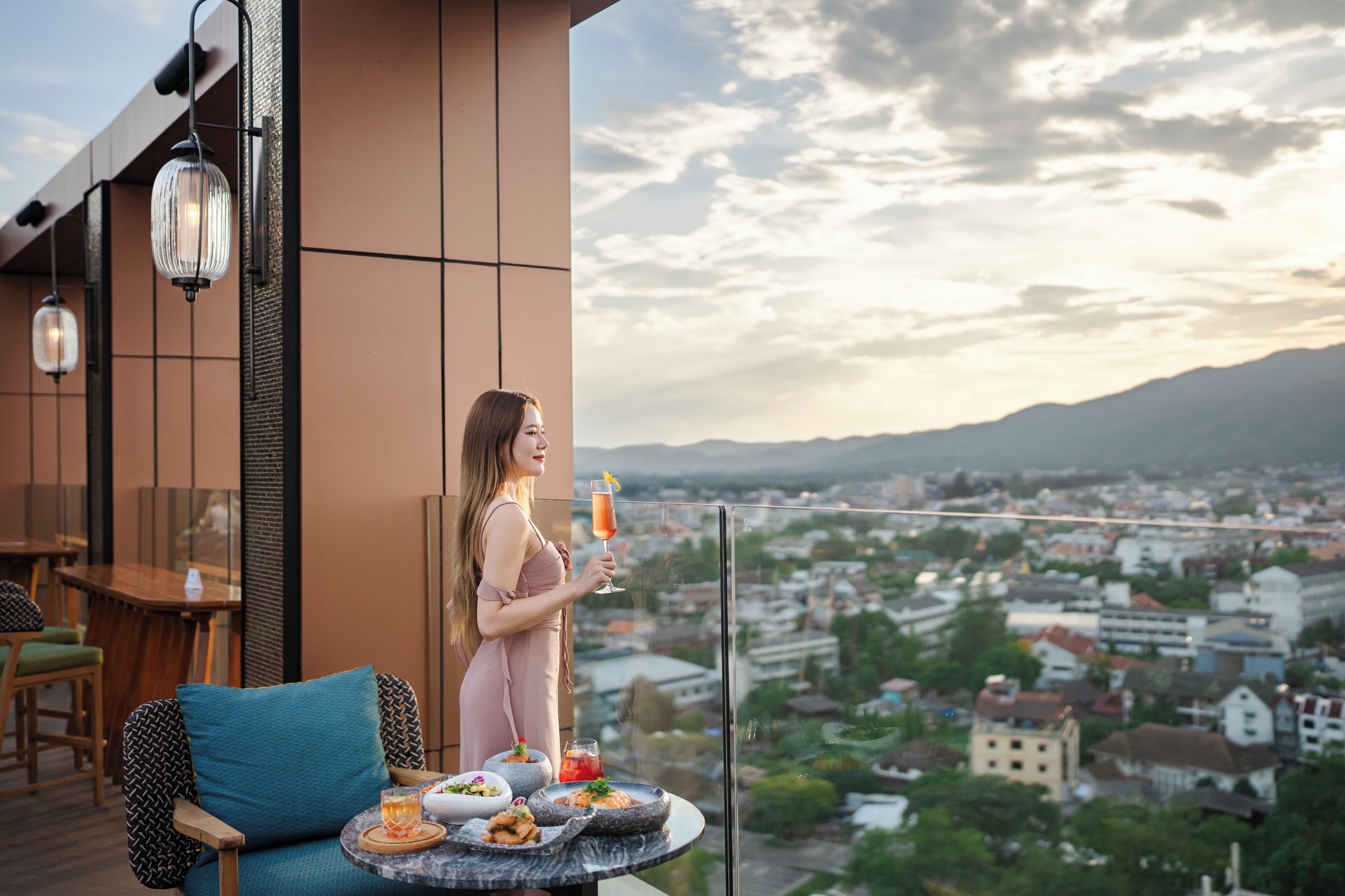 Une femme se tient sur un balcon, avec vue sur la ville, et tient dans ses mains une boisson et une assiette de nourriture.