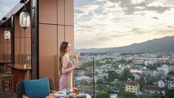 Une femme se tient sur un balcon, avec vue sur la ville, et tient dans ses mains une boisson et une assiette de nourriture.
