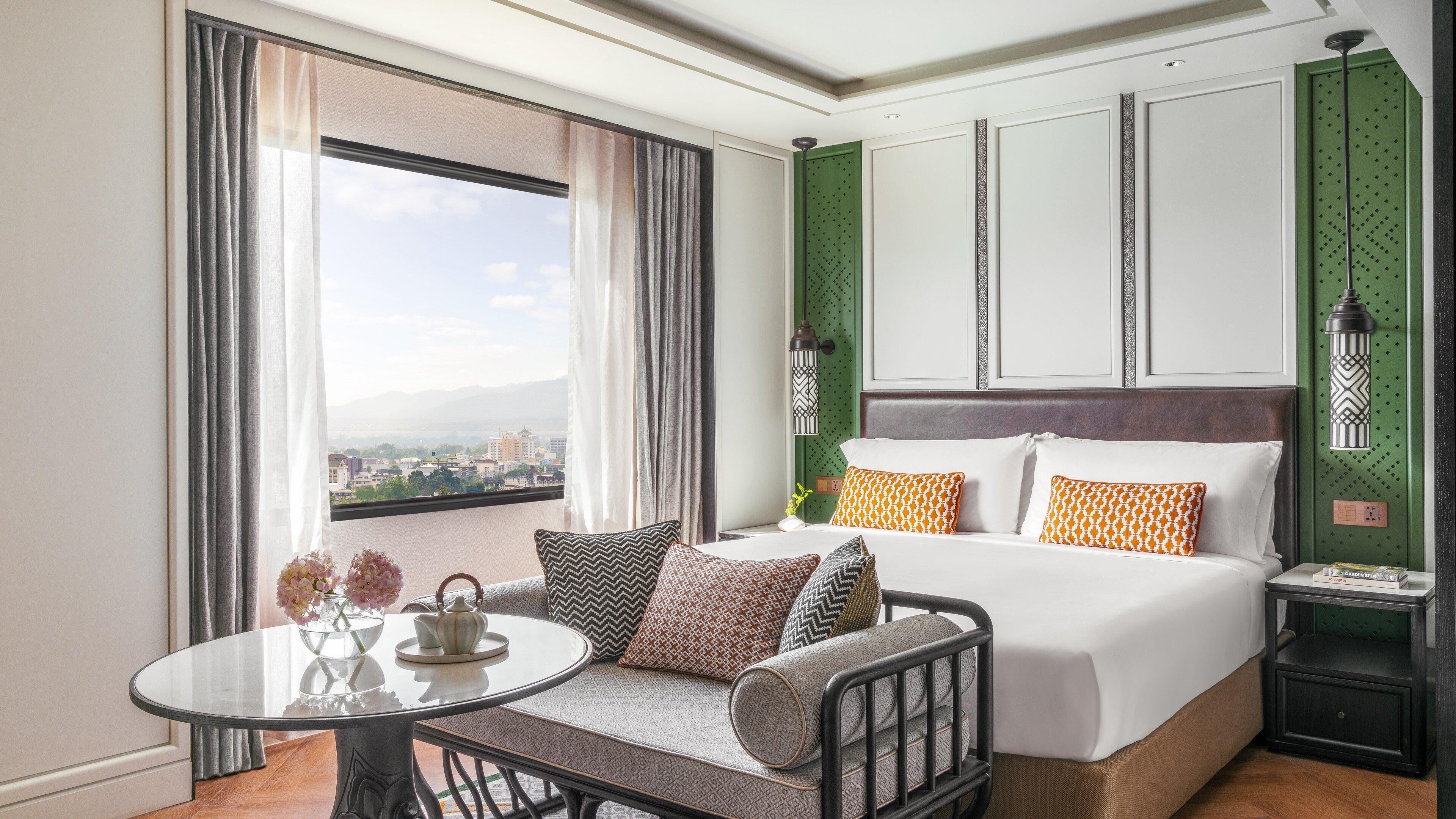 A hotel room with a bed, a table with a vase and teapot, a view of the city, and a green accent wall.