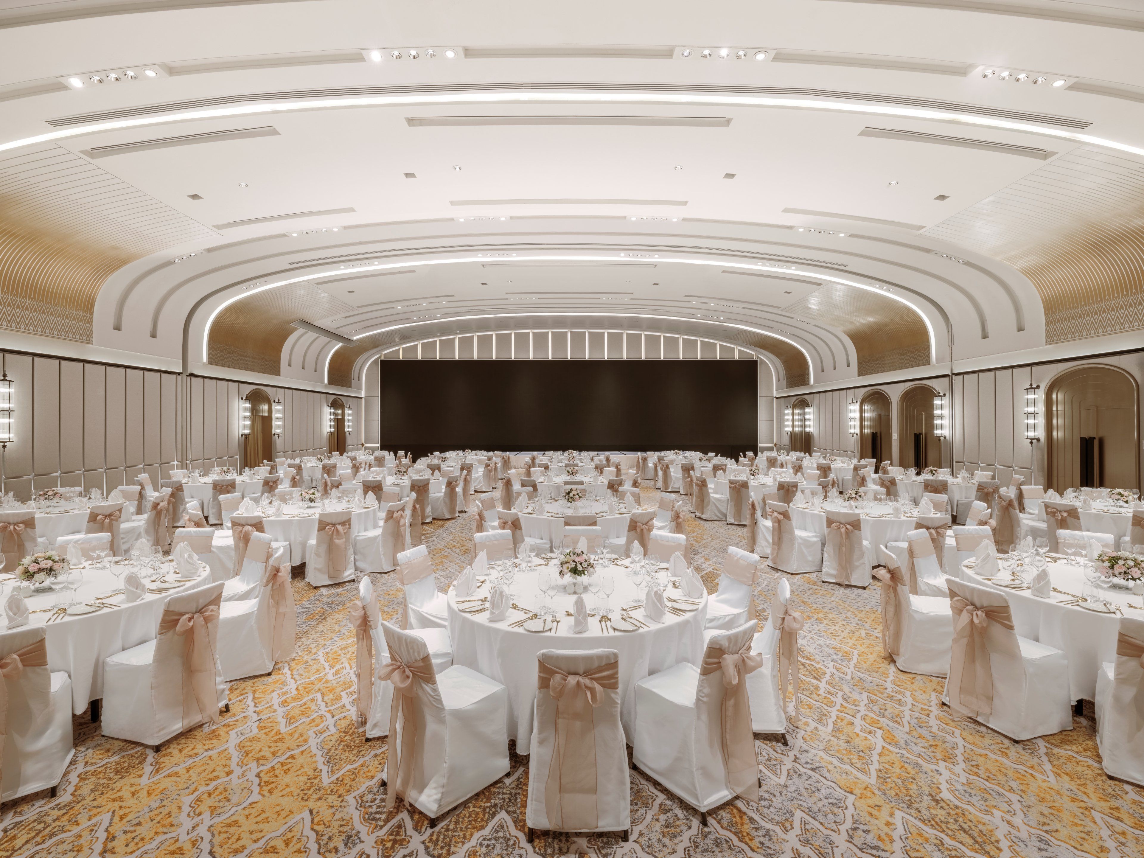 Un amplio salón de banquetes con mesas redondas, manteles blancos y sillas con lazos color beige.