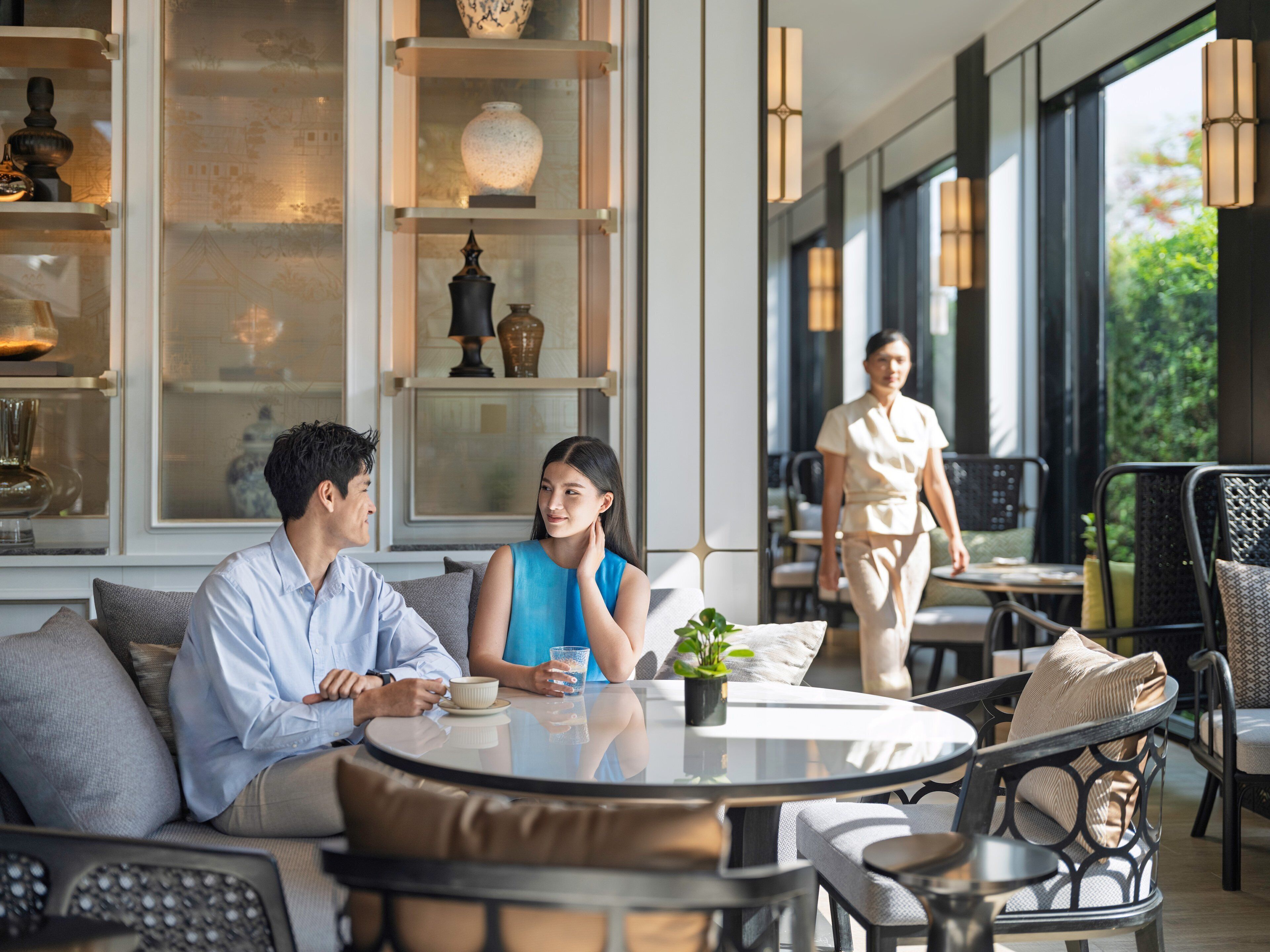 A modern interior with a round glass table, comfortable seating, and a large window offering a view of greenery.